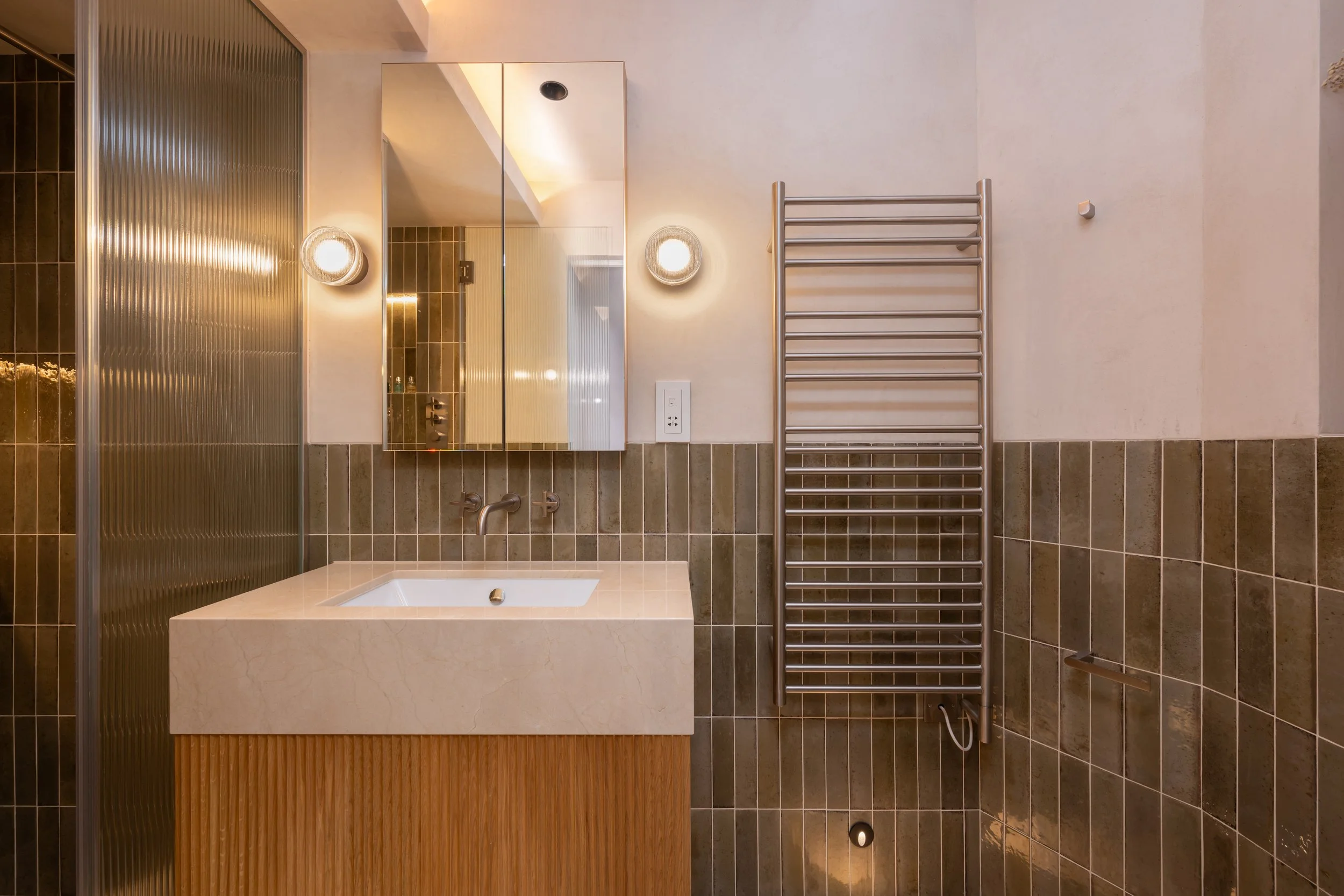Modern bathroom with a sink, mirrored cabinet, wall-mounted lights, radiator, brown tiled walls, and a textured glass shower door.