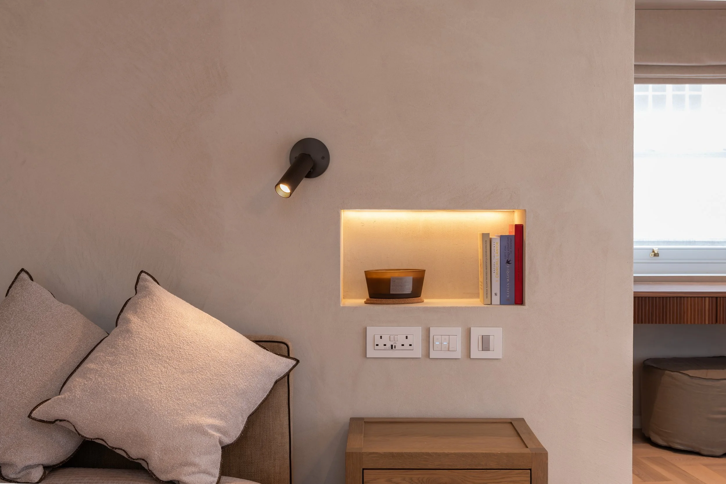 Interior of a bedroom with a beige wall, black wall-mounted lamp, built-in shelf with books and decorative bowl, electrical outlets, and a wooden side table. part of a bed with beige pillows and a window with blinds is visible.