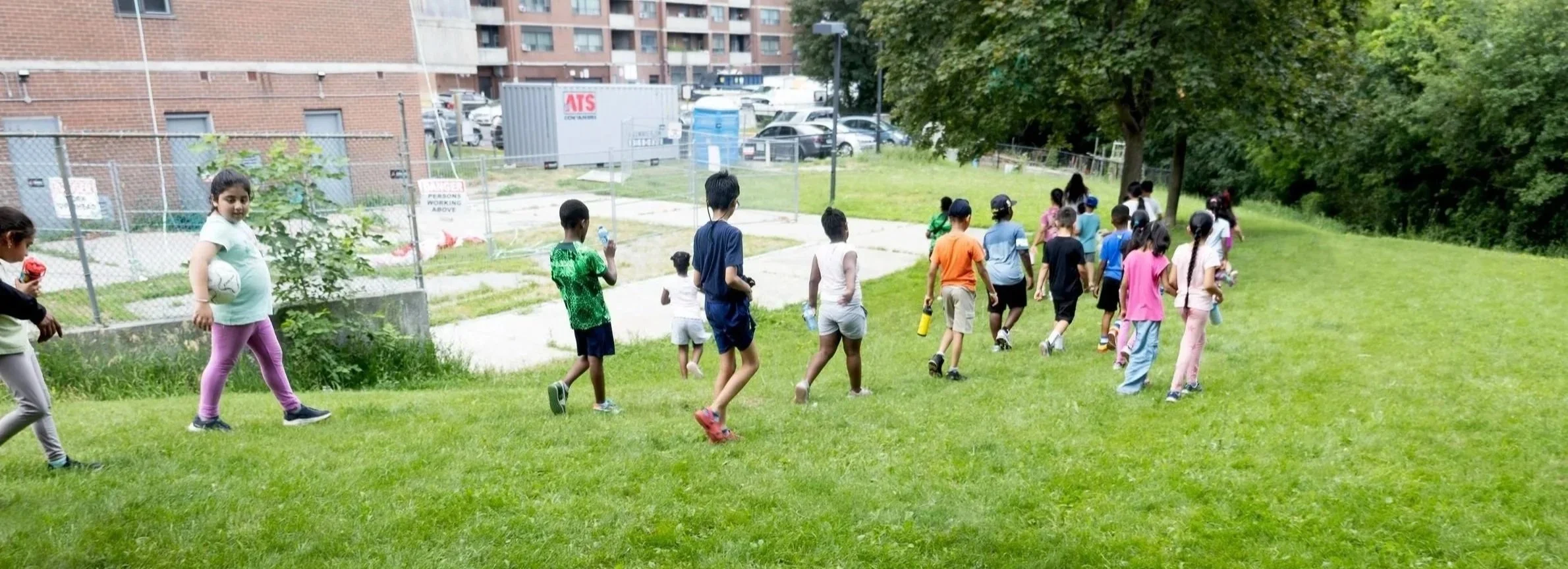20 children in the RWC summer camp walking through a grassy field. One child is holding a soccer ball.