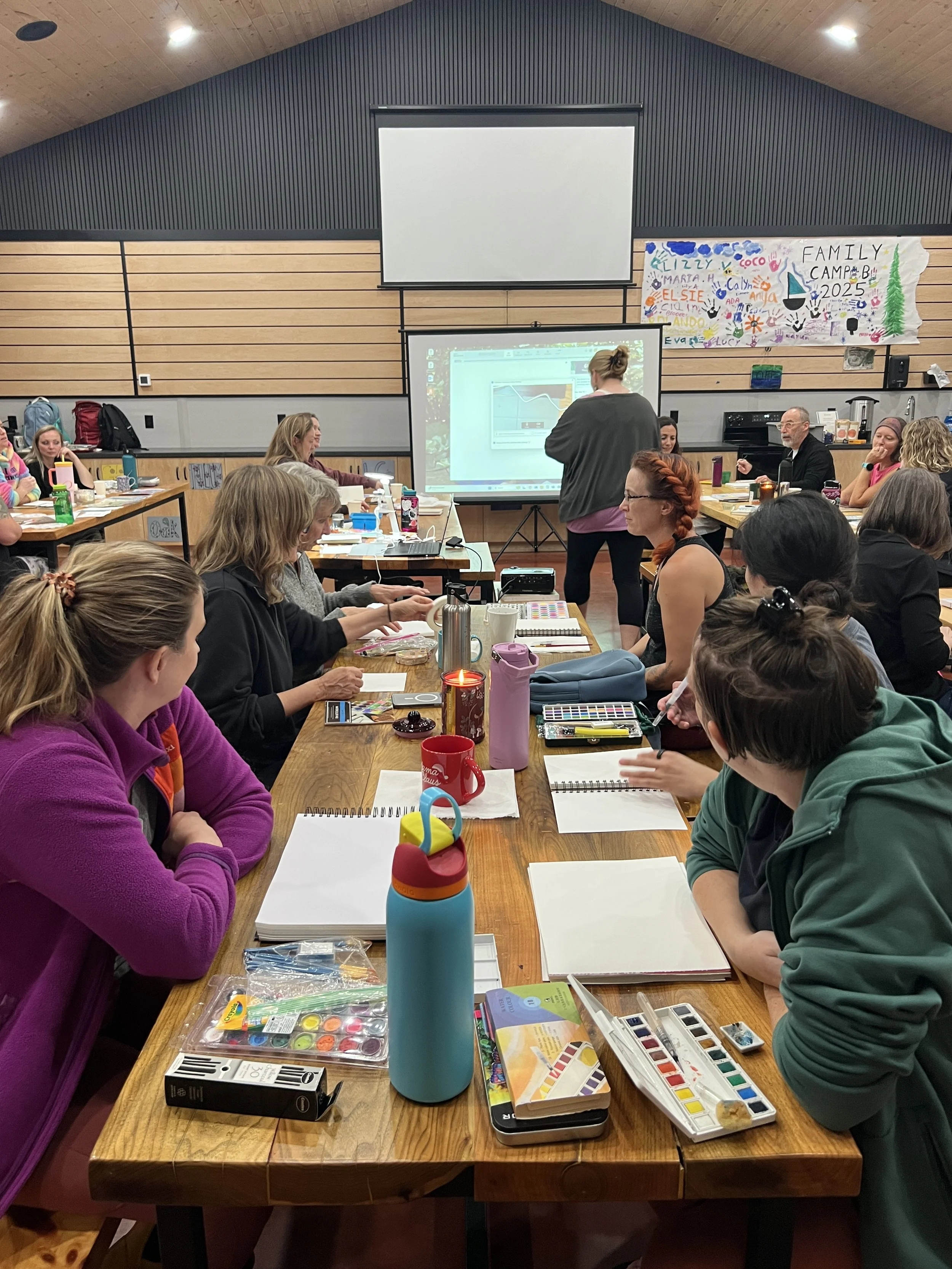 Two women sitting at a wooden table, writing on papers, with various items including a mug, water bottle, and snacks in front of them.