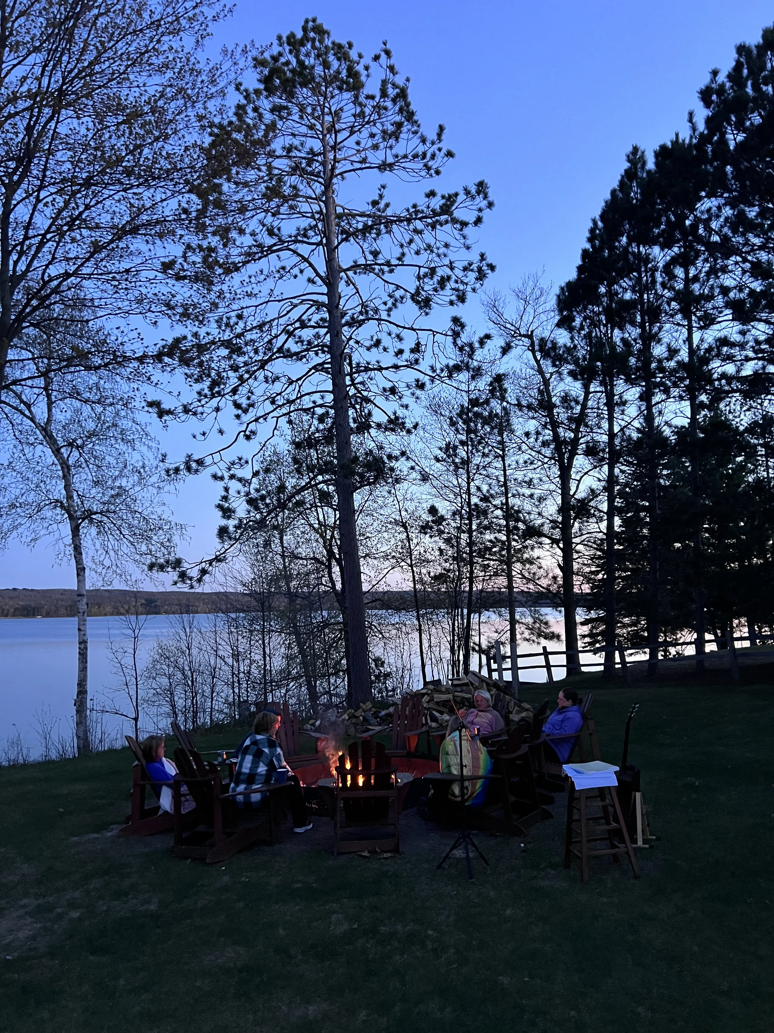 People sitting around a campfire outdoors at dusk with trees and a lake in the background.