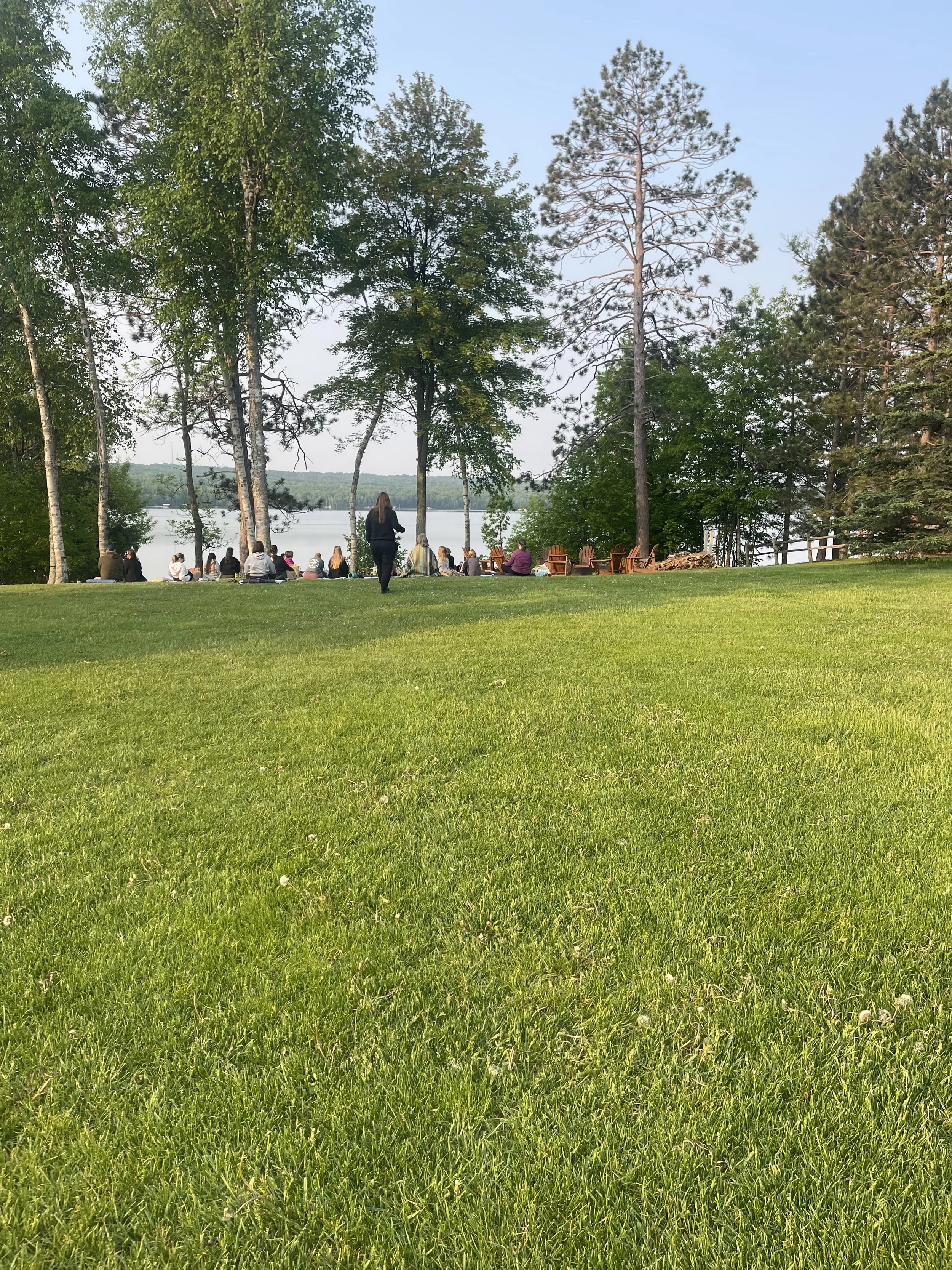 A group of people sitting and standing on a grassy area near a lake, surrounded by tall trees, with a clear sky above.