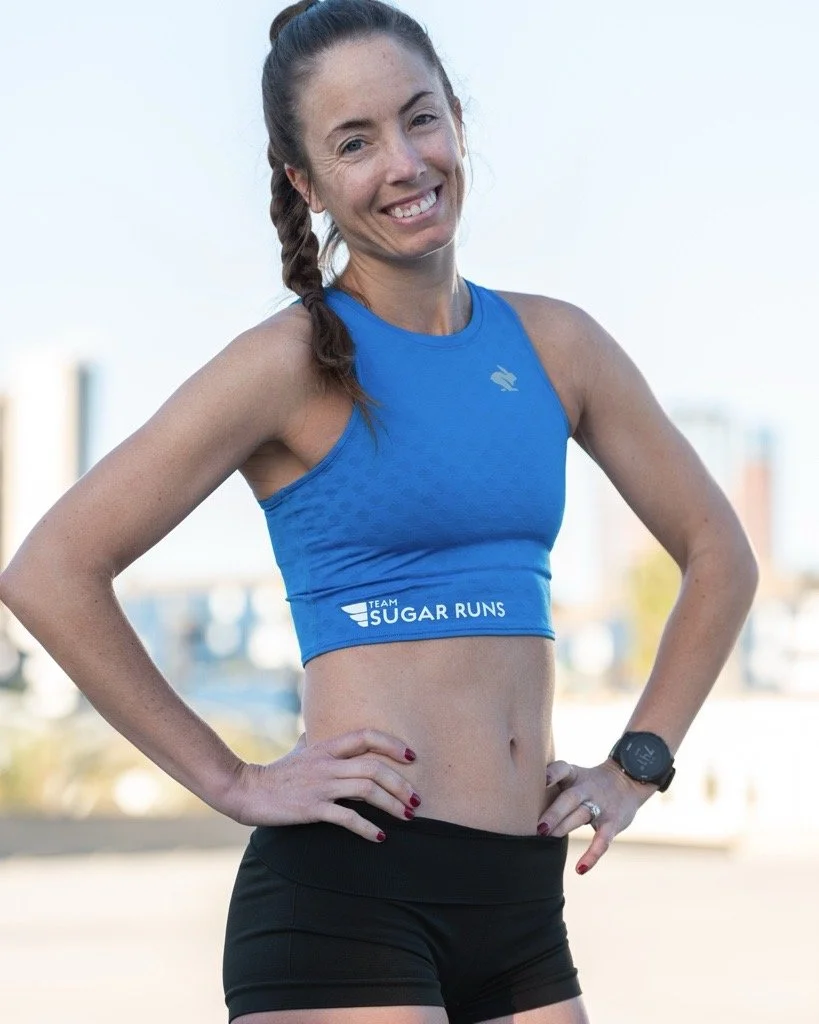A woman in a blue crop top and black shorts smiles outdoors with hands on hips, wearing a watch and a braid.