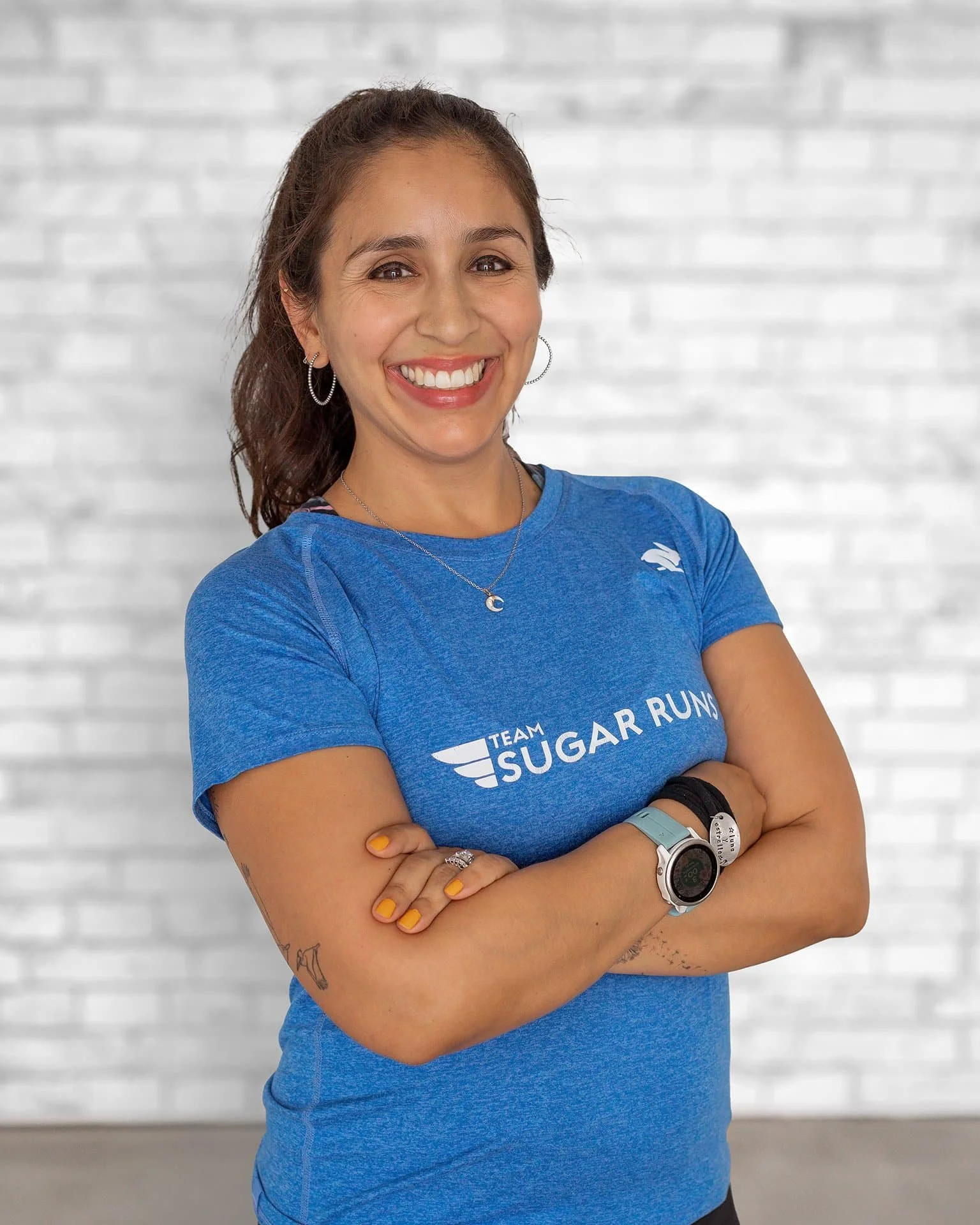 A woman in a blue shirt smiles against a brick wall with arms crossed, wearing a watch and ponytail