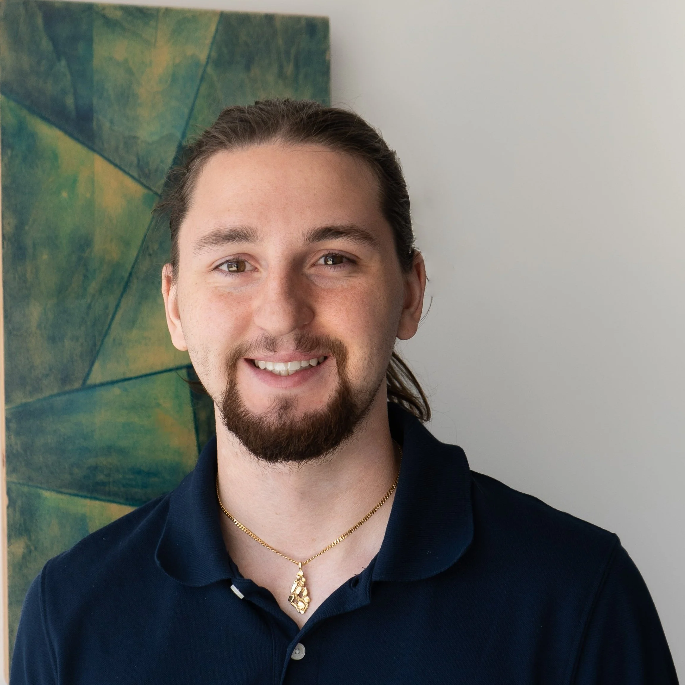 A smiling young man with long hair and a goatee, wearing a navy blue polo shirt and a gold necklace, standing next to a green and yellow abstract painting.