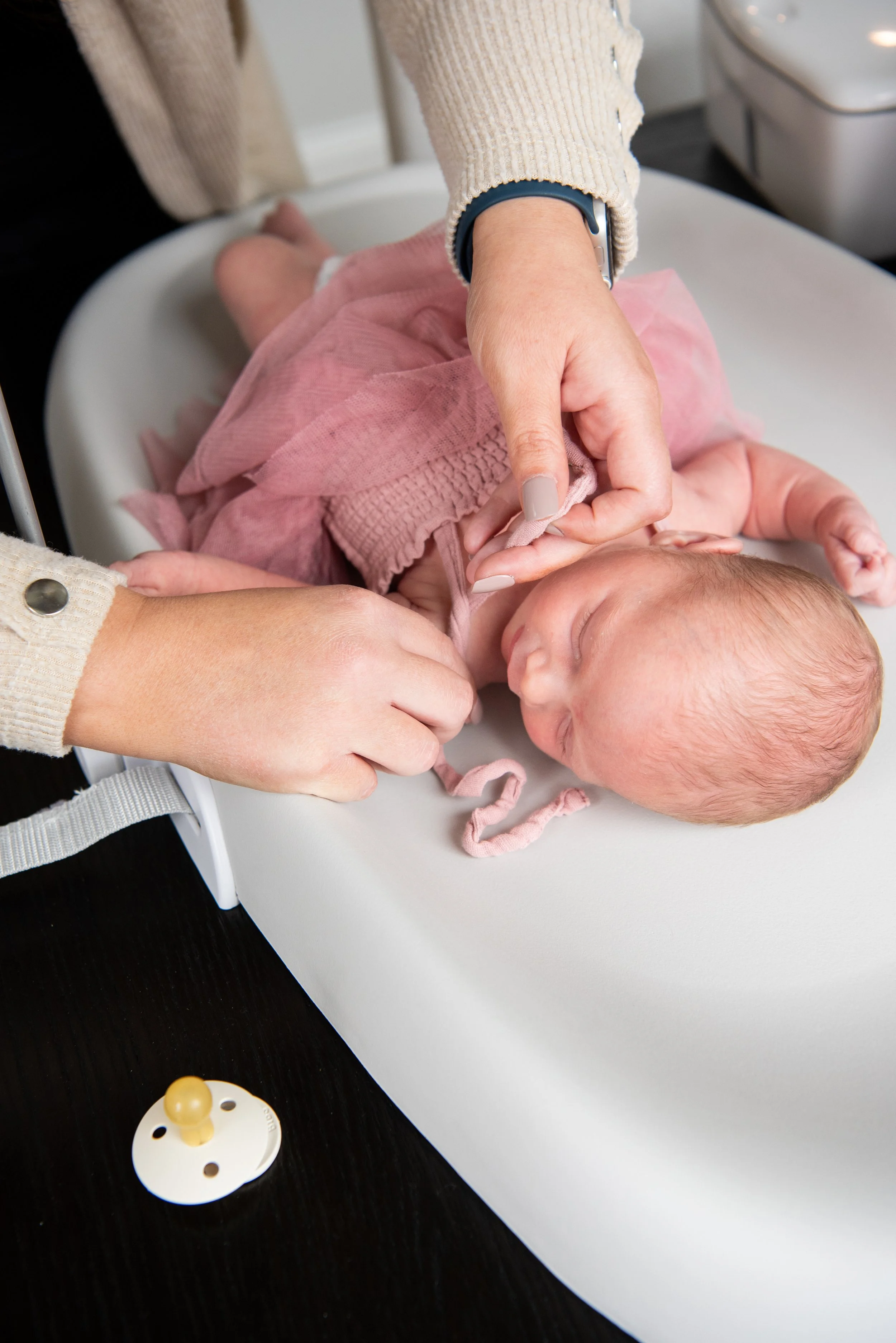 Newborn baby girl in a pink dress with hands tying a bow at the shoulder