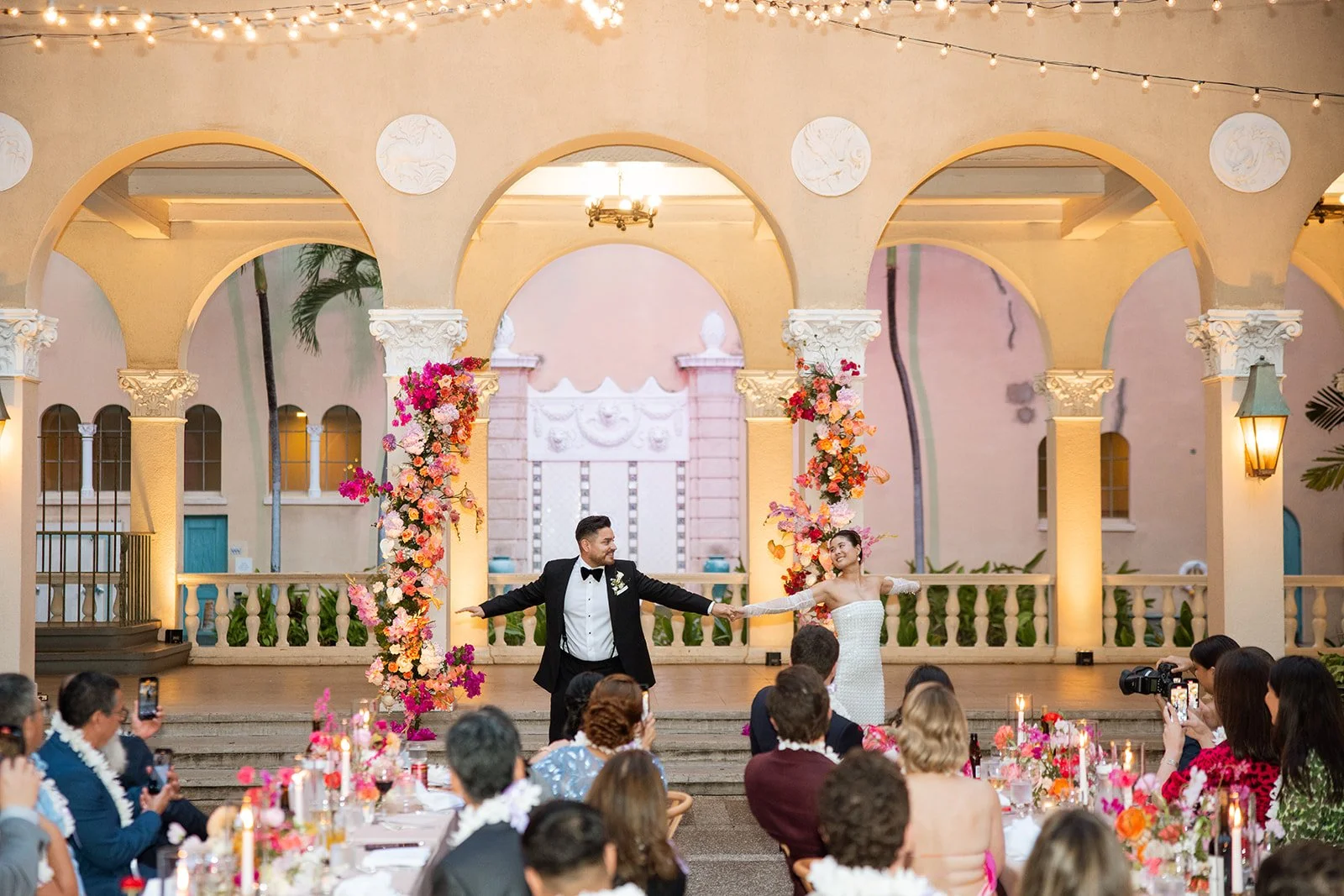 A bride and groom are dancing at their wedding reception. They are holding hands and smiling, with guests seated at tables decorated with flowers and candles watching and taking photos. The background features an archway decorated with colorful flowers, and string lights hanging above.