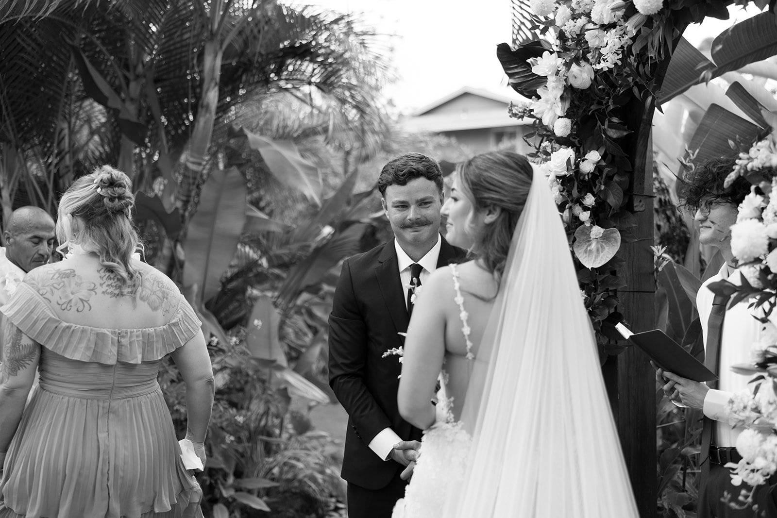 Black and white photo of a wedding ceremony with a groom and bride holding hands, standing under a floral arch, surrounded by officiant and guests, outdoors among lush tropical plants.