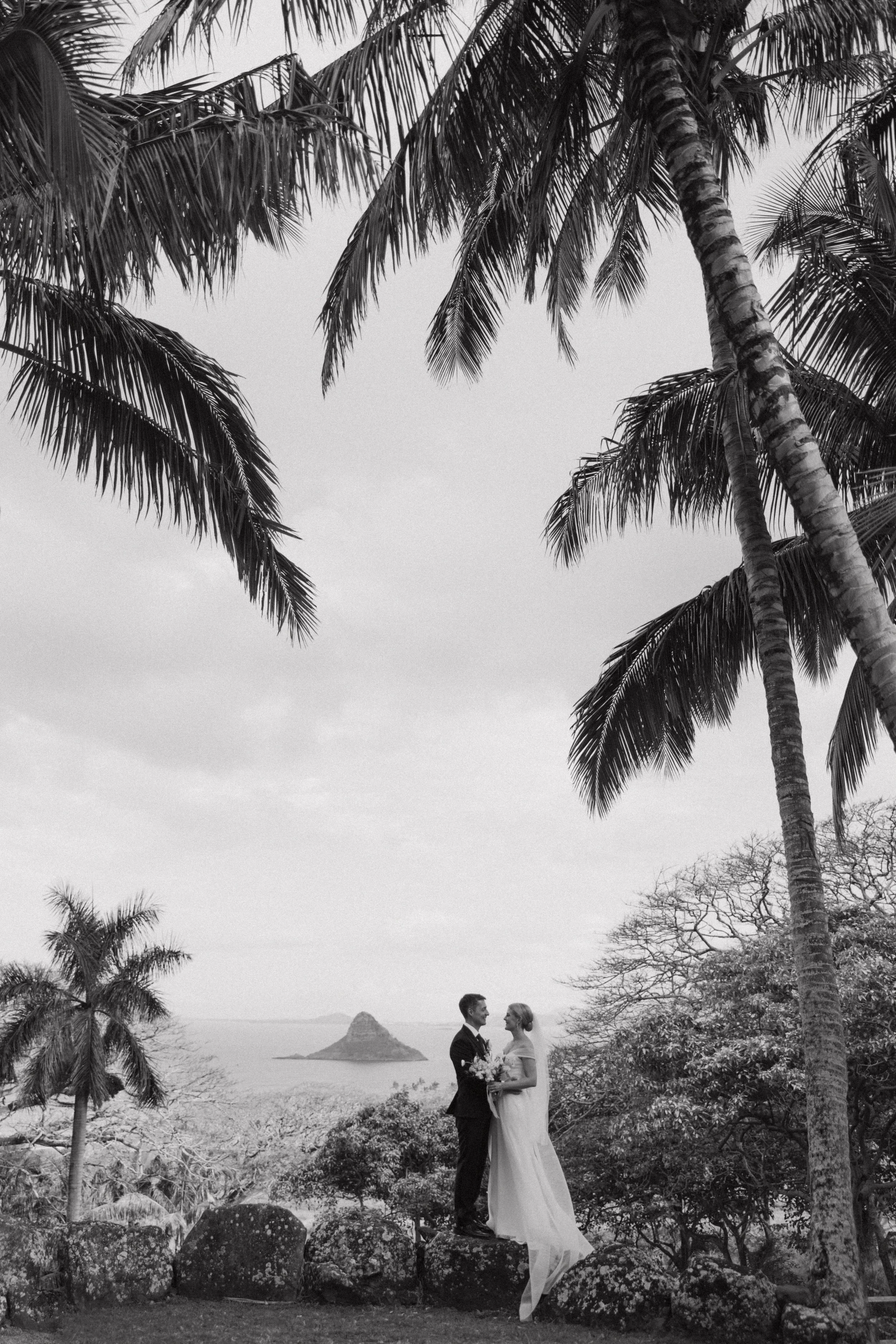 A black and white photo of a bride and groom standing on rocks among trees in Hawaii for their wedding day, with palm trees in the foreground and an island in the distance.