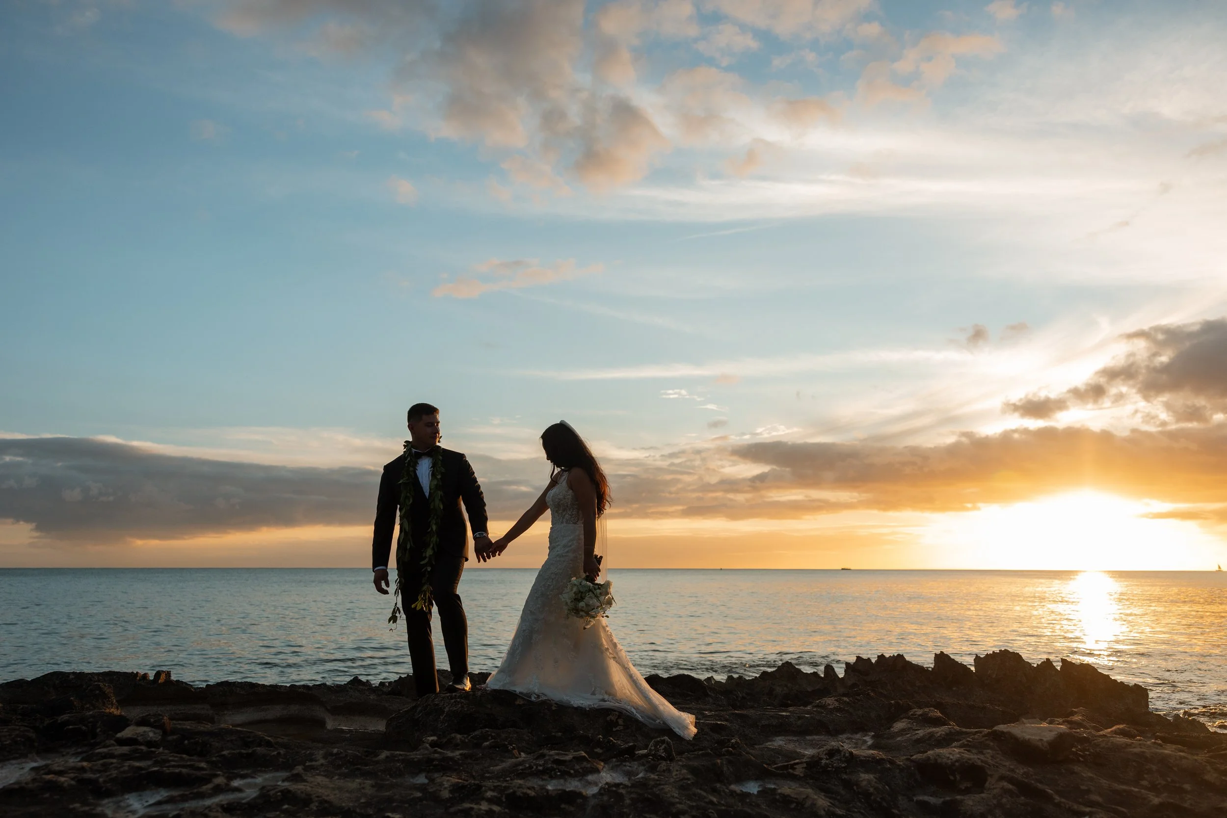 A bride and groom holding hands on a rocky Hawaiian shoreline at sunset, with the ocean and sky in the background.