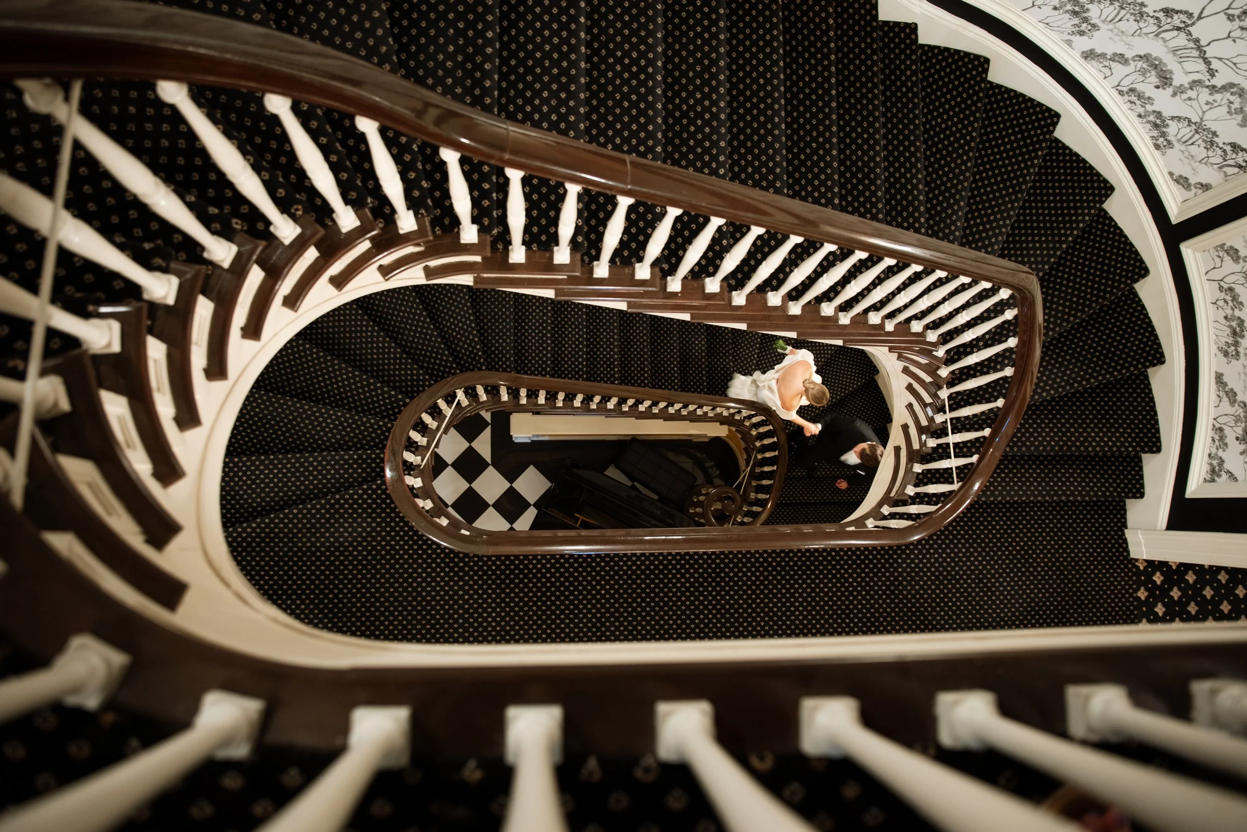 Top-down view of a spiral staircase with a black and white patterned carpet, wooden handrails, and two newlyweds standing on one of the intermediate landings, one in a white wedding dress holding a bouquet.