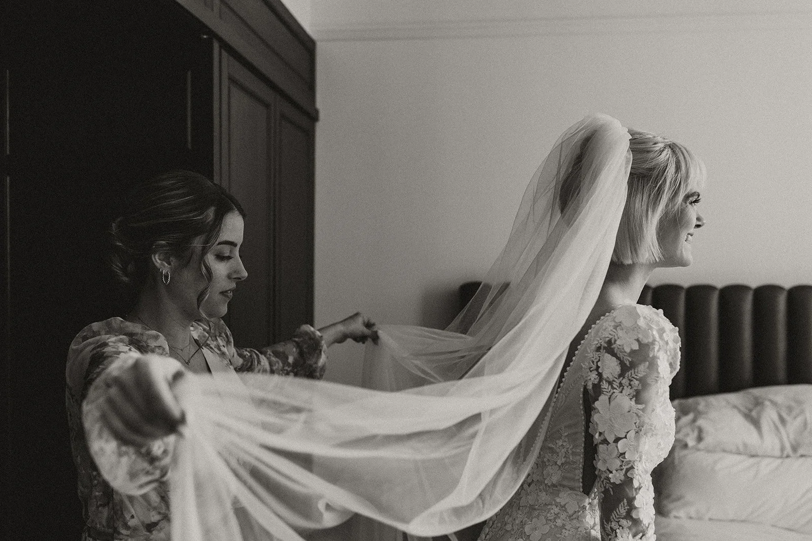 A bride getting ready for her wedding day with the help of her bridesmaid , who is adjusting her cathedral length veil in a luxury hotel suite.
