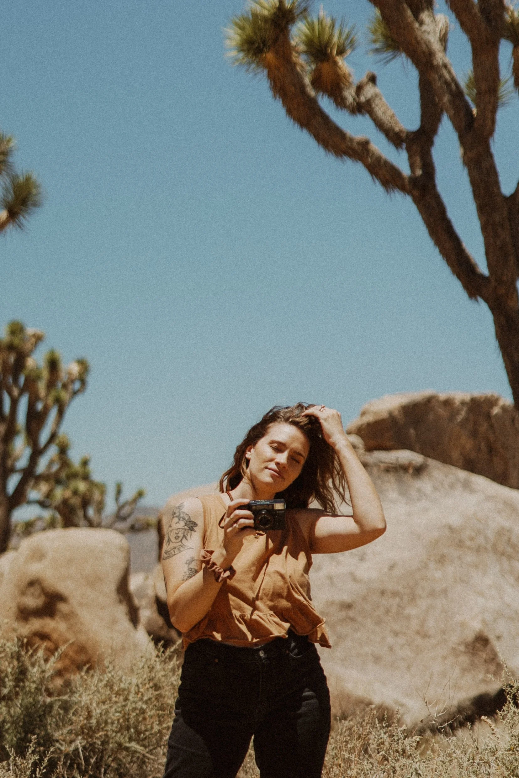 Grace Troutman holding a camera in a desert landscape with rocks and Joshua trees under a clear blue sky.