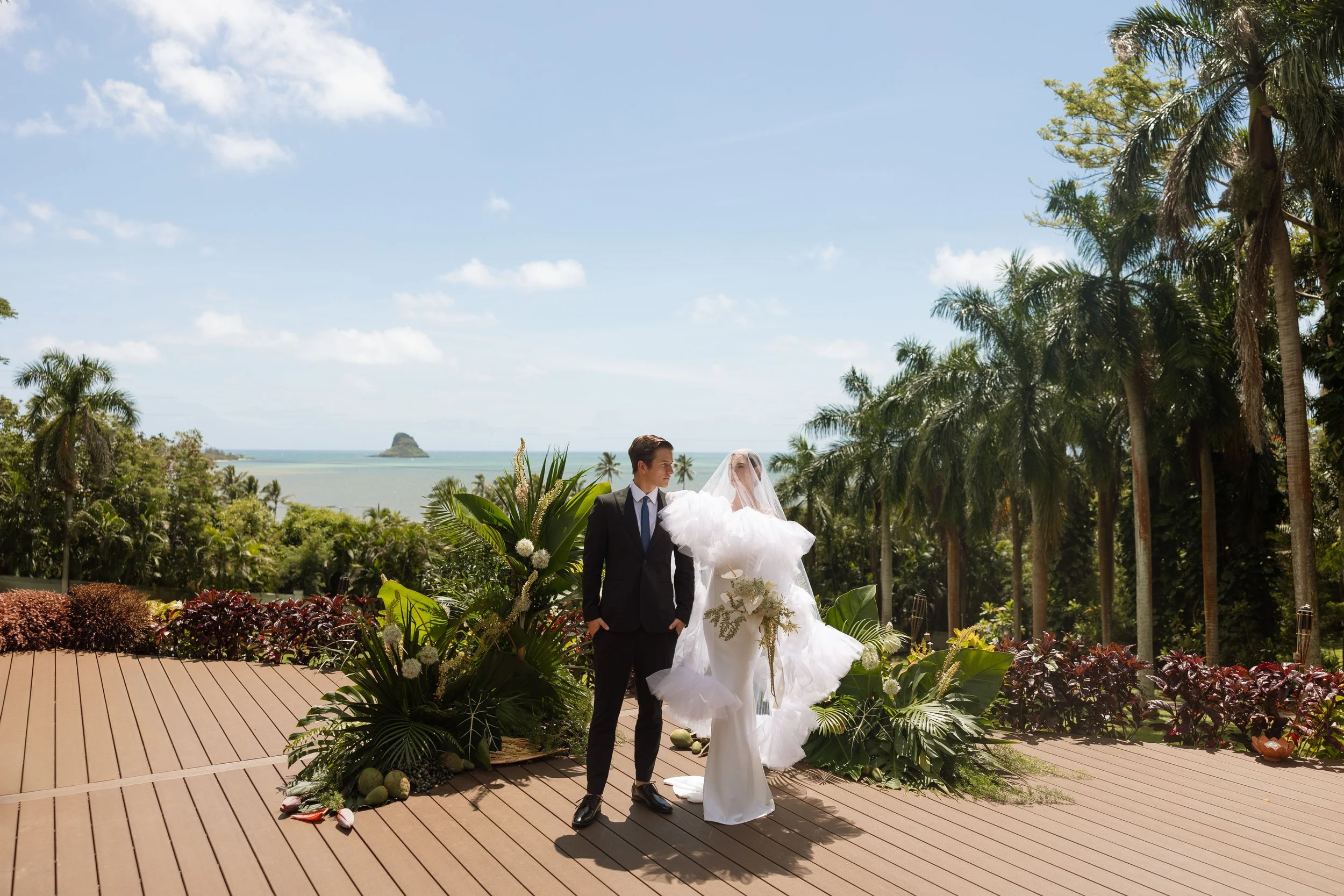 A bride and groom in Hawaii standing outdoors on a wooden platform, surrounded by lush green tropical plants and palm trees, with a view of the ocean and a small island in the background under a partly cloudy sky.