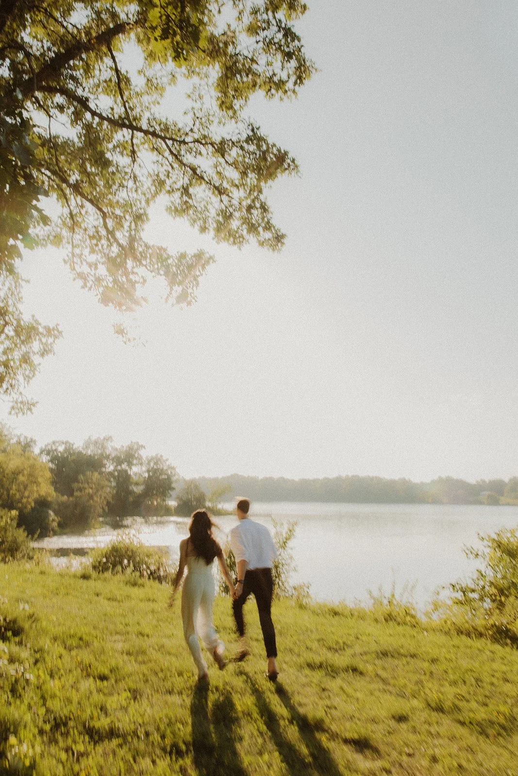 A couple walking hand in hand along a grassy shore near a lake during sunset, surrounded by trees.