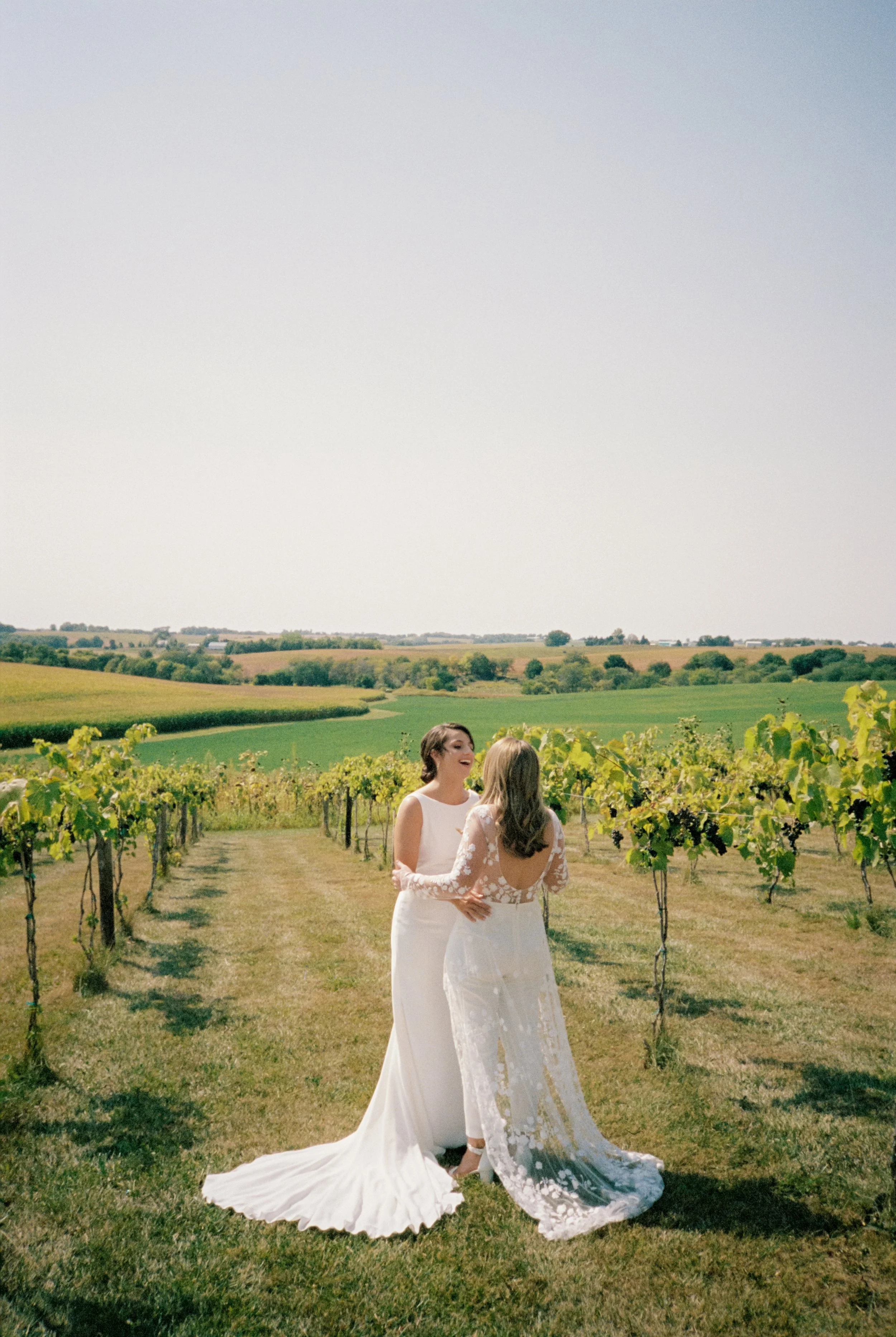 Two women in wedding dresses standing in a vineyard, smiling and talking together, with green hills and a clear sky in the background.