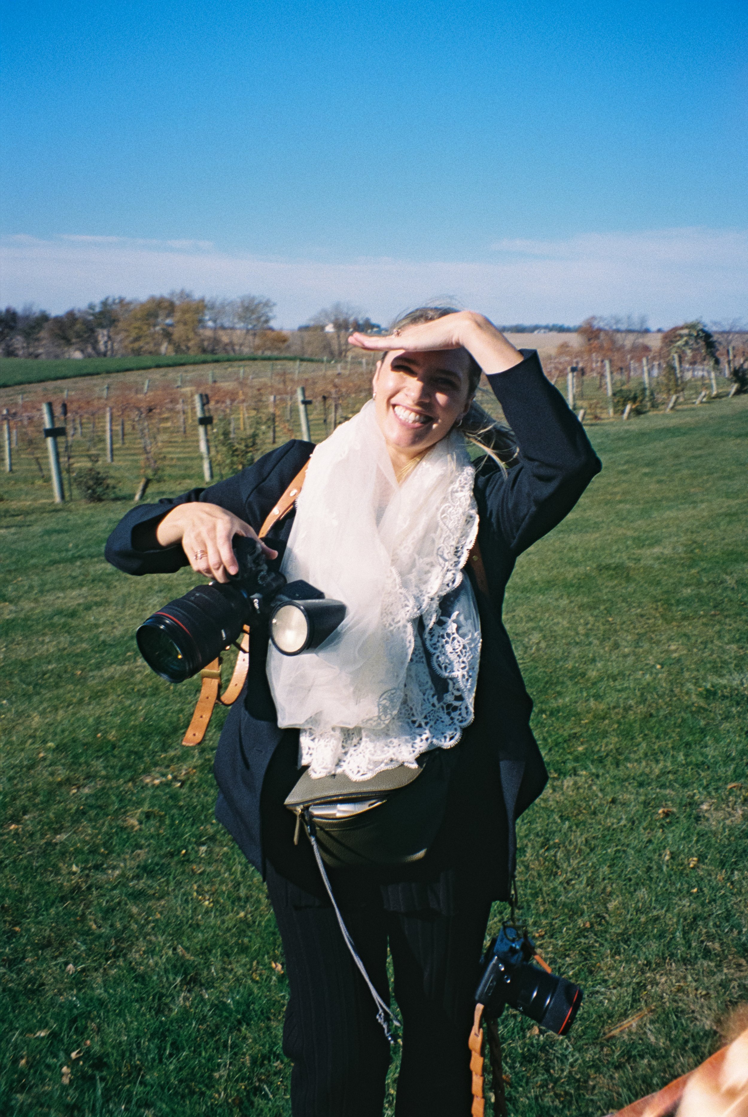 A woman outdoors on a sunny day, smiling and using her hand to shield her eyes from the sun, holding a professional camera with a large lens, with a scenic background of green grass, trees, and a clear blue sky.