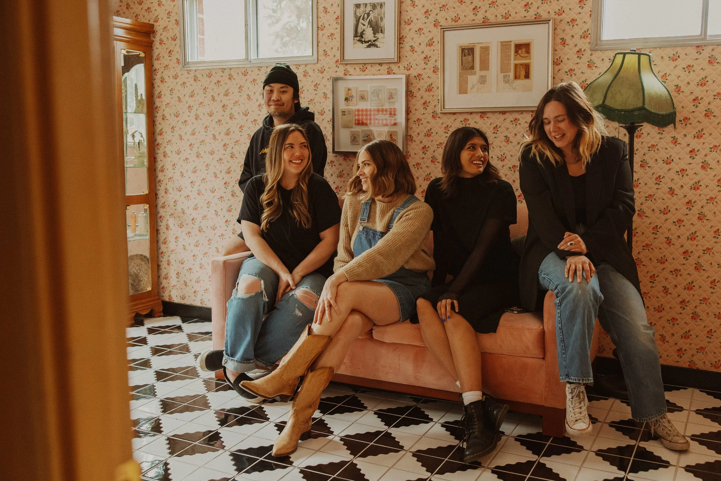 Grace Troutman's photography team sitting and standing on a pink couch in a cozy, vintage-style room, smiling and engaging in conversation.