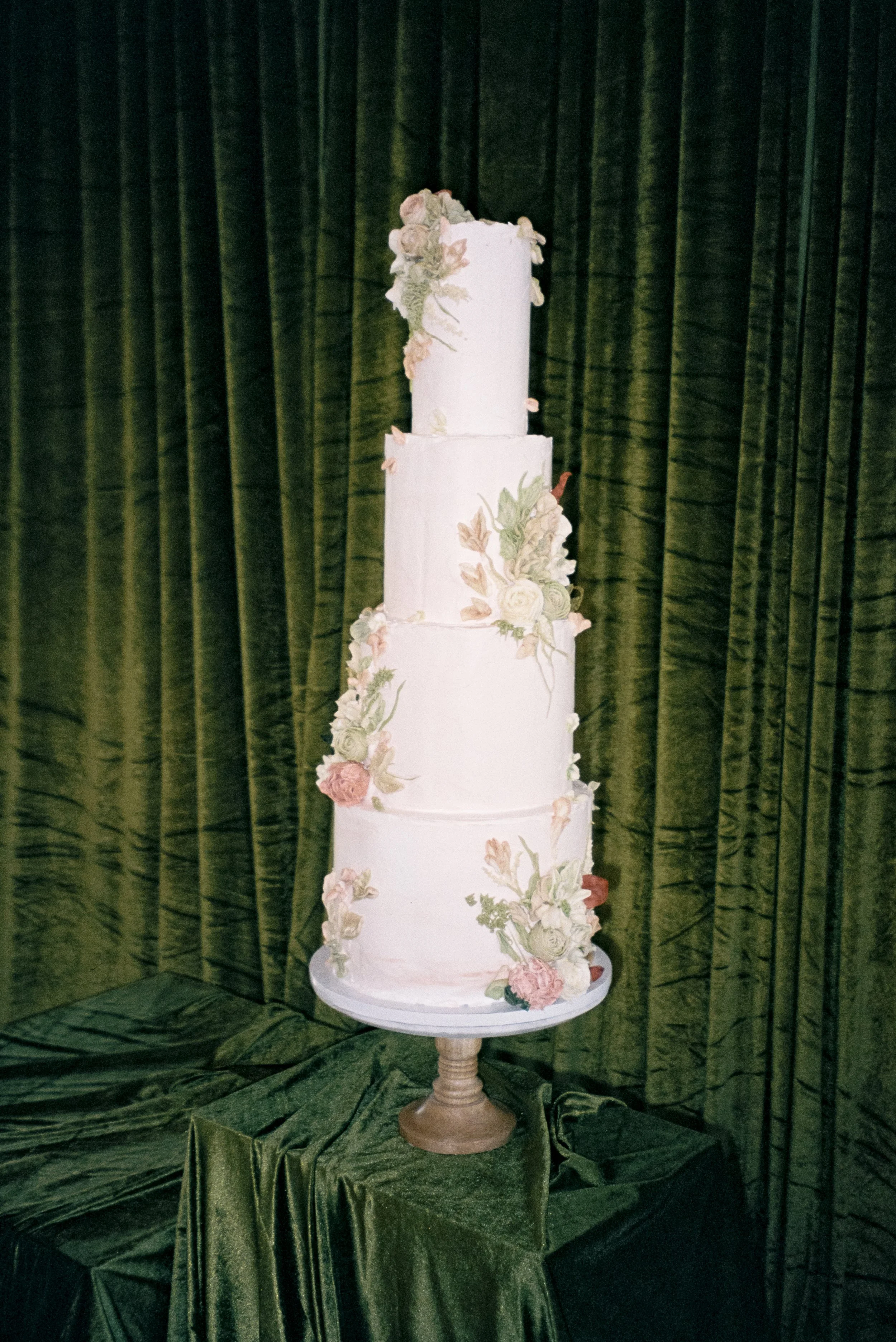 A tall 4-tier white wedding cake with floral decorations on a wooden stand, set against green velvet curtains.