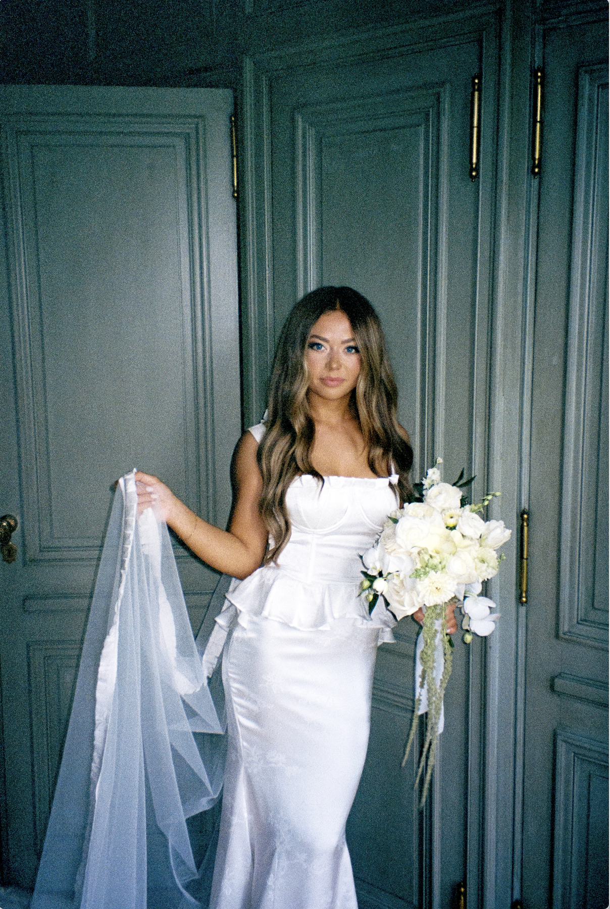 A woman in a white wedding dress holding a bouquet of white flowers, standing in front of a textured teal door in a historic mansion.