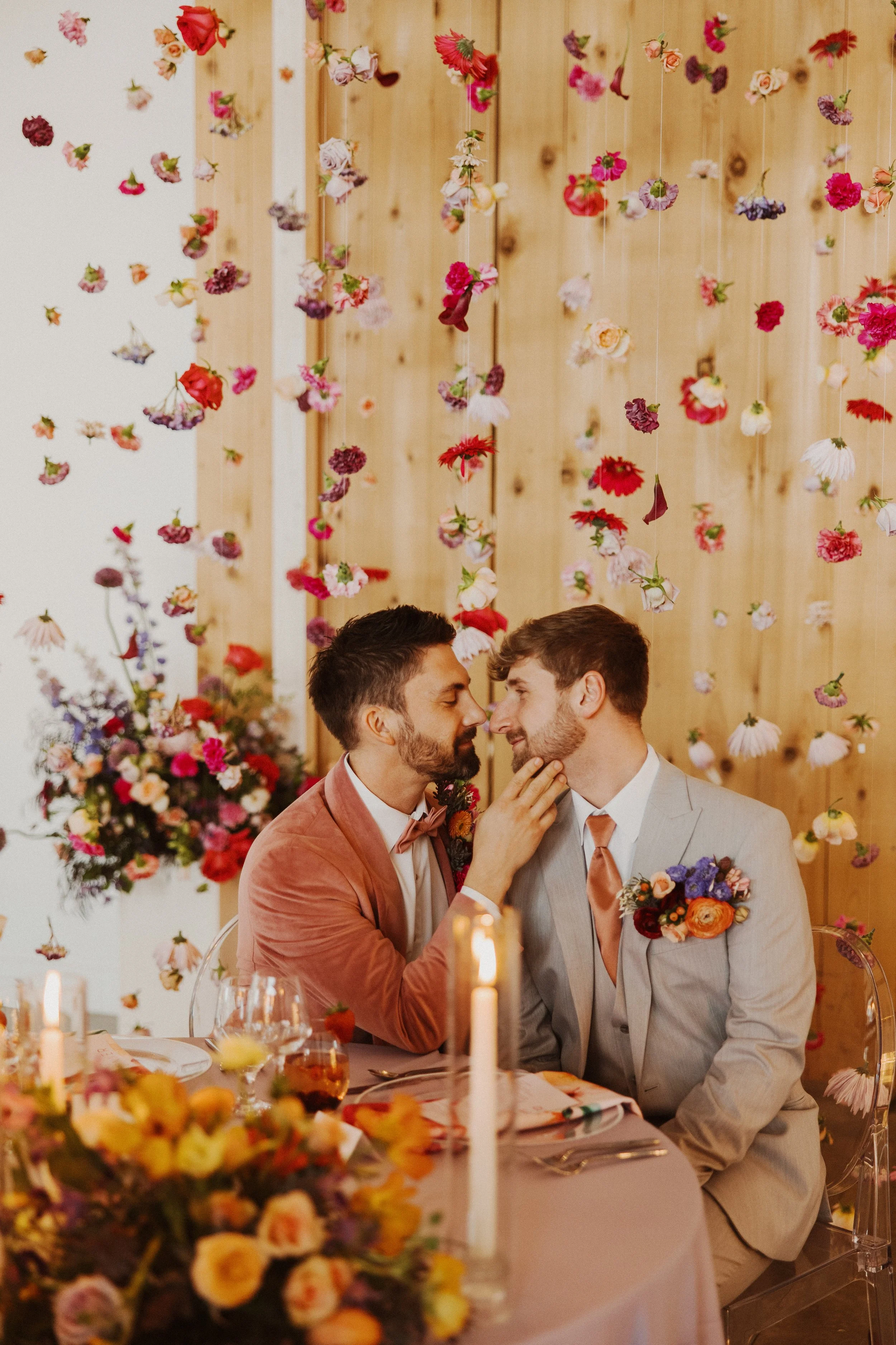A gay couple in suits sharing an intimate moment at their wedding reception, surrounded by floral decorations and hanging flowers.