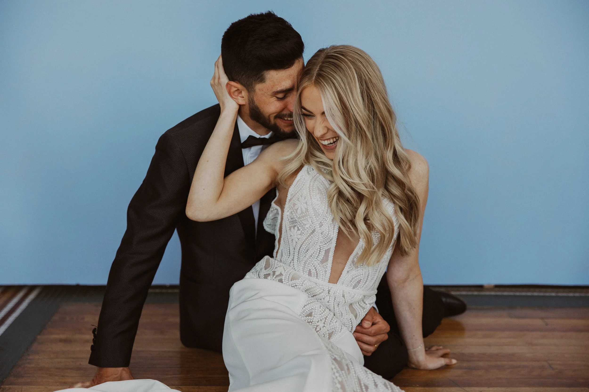 A man and woman in formal attire sharing an intimate and joyful moment, with the man in a tuxedo and the woman in a white lace dress, sitting on a wooden floor against a light blue wall.