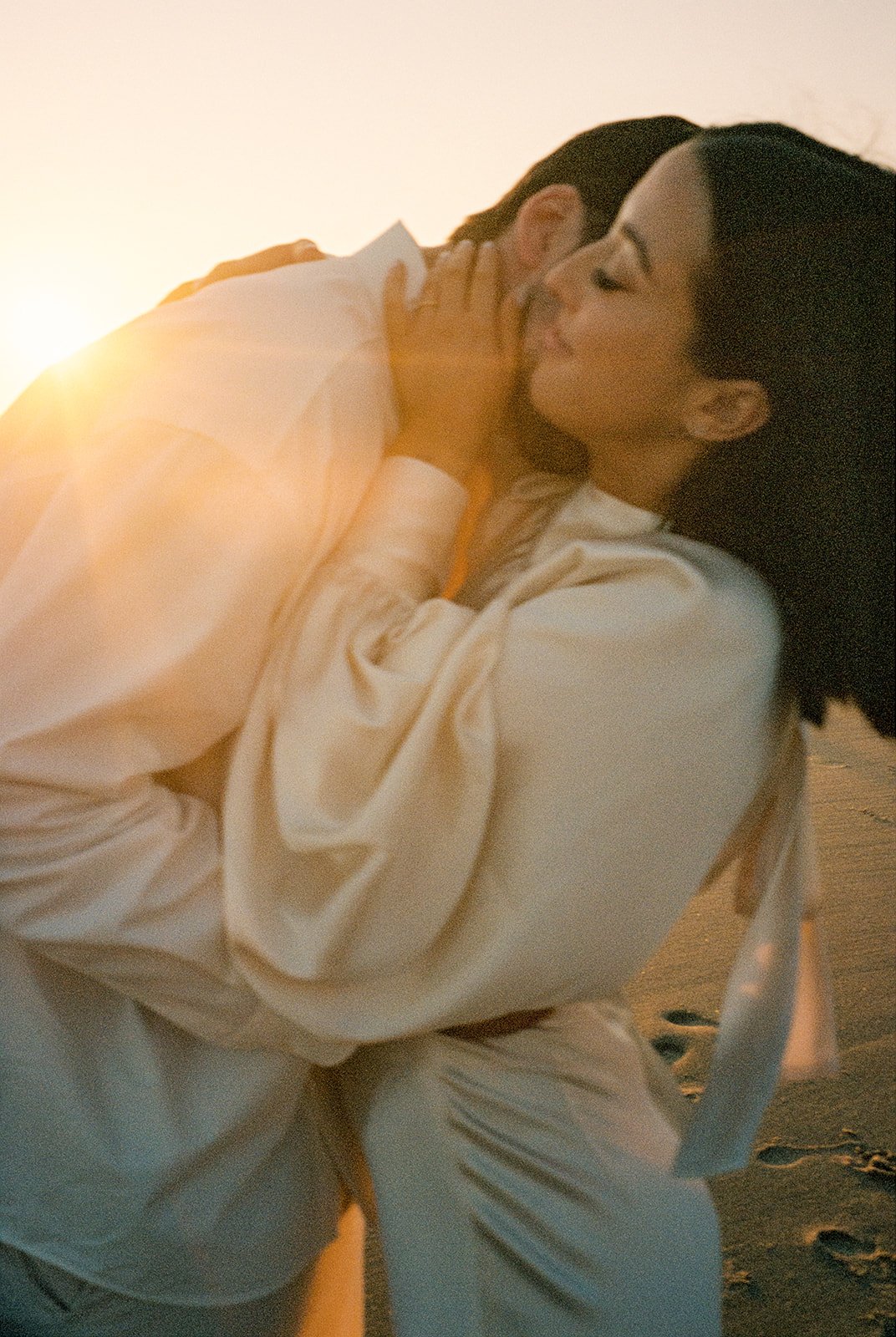 A couple embracing on the beach at sunset, with the man holding the woman close and the woman closing her eyes