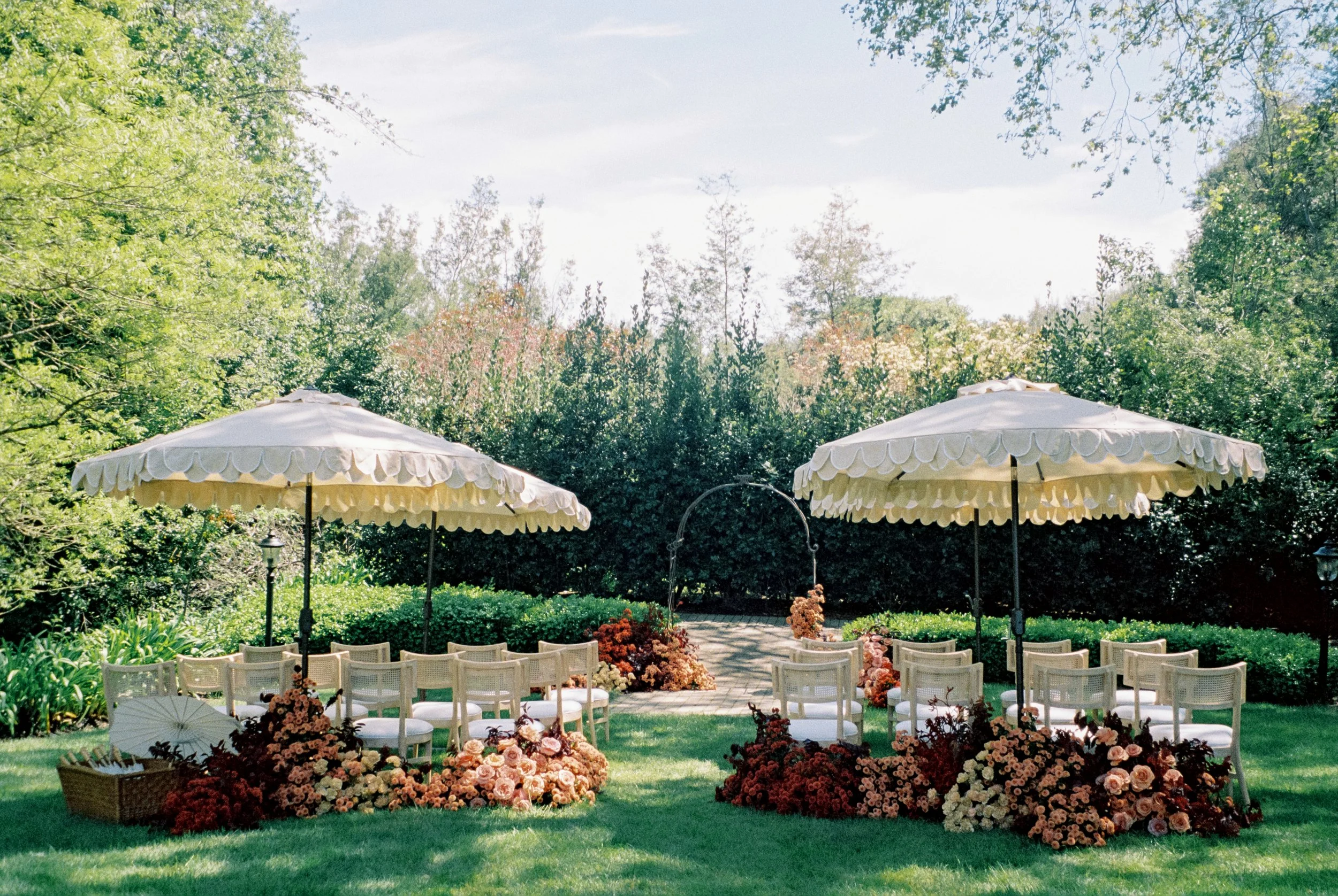 An outdoor wedding ceremony setup with two large white umbrellas, white chairs arranged in rows, floral arrangements on the ground, and an arch in the background, all surrounded by green trees and bushes at the Madrona in California.