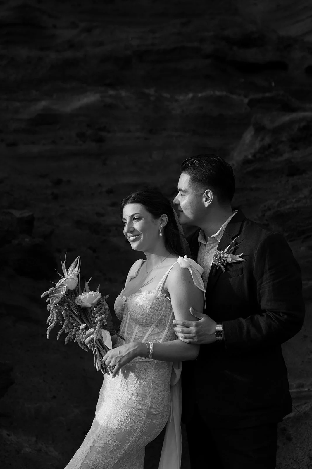A black and white photograph of a bride and groom standing close together, with the groom kissing the bride's temple. The bride is smiling and holding a bouquet of flowers, wearing a lace wedding dress with shoulder bows. The groom is in a dark suit with a boutonniere, smiling with his eyes closed.