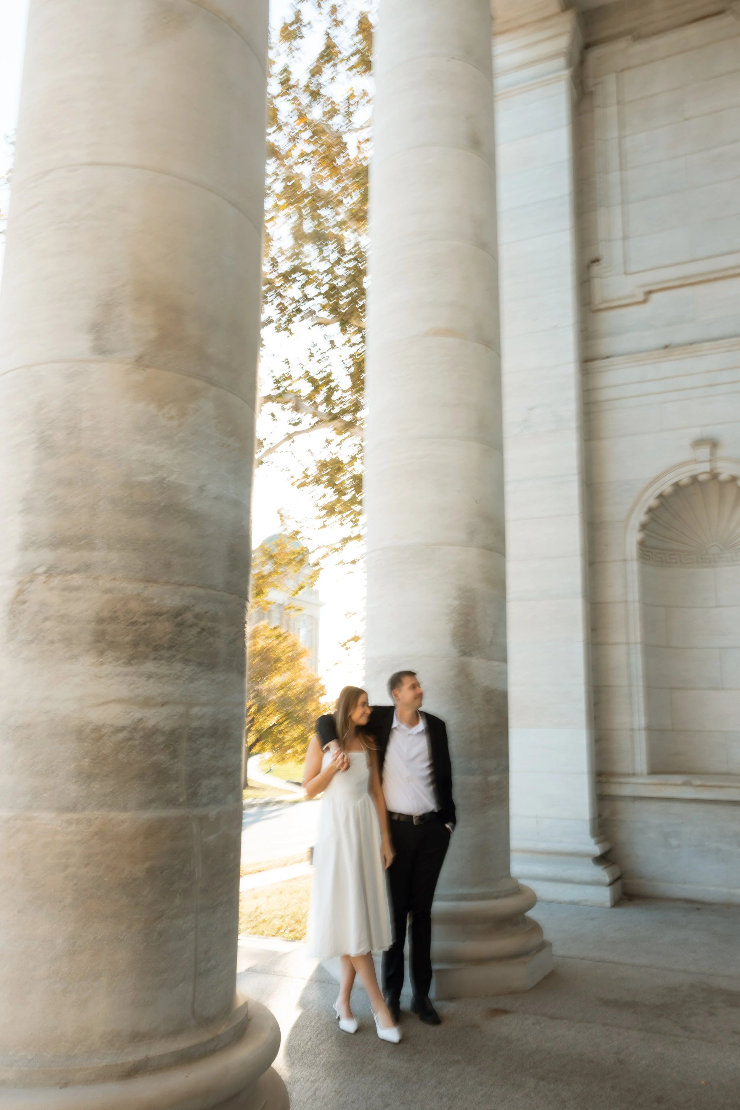 A couple stands beneath large stone columns near a historic building, with autumn trees in the background.