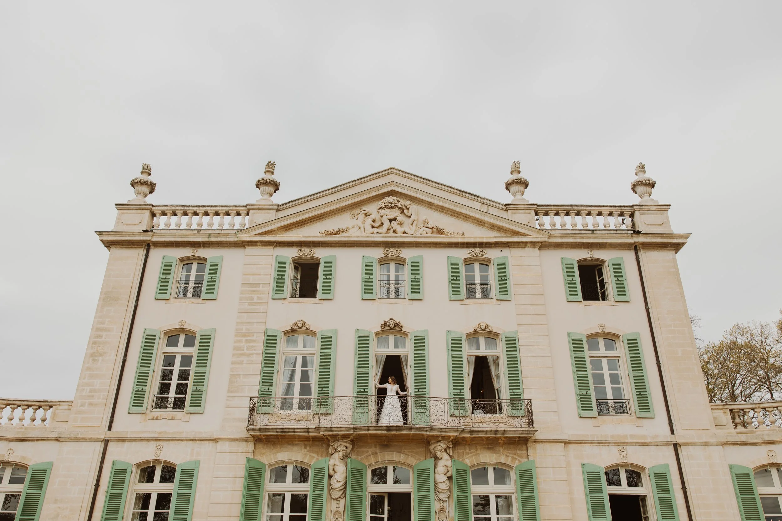 A grand, historic chateau in France with multiple windows, green shutters, decorative stone carvings, and a balcony with a bride standing on it, looking out looking elegant and beautiful.