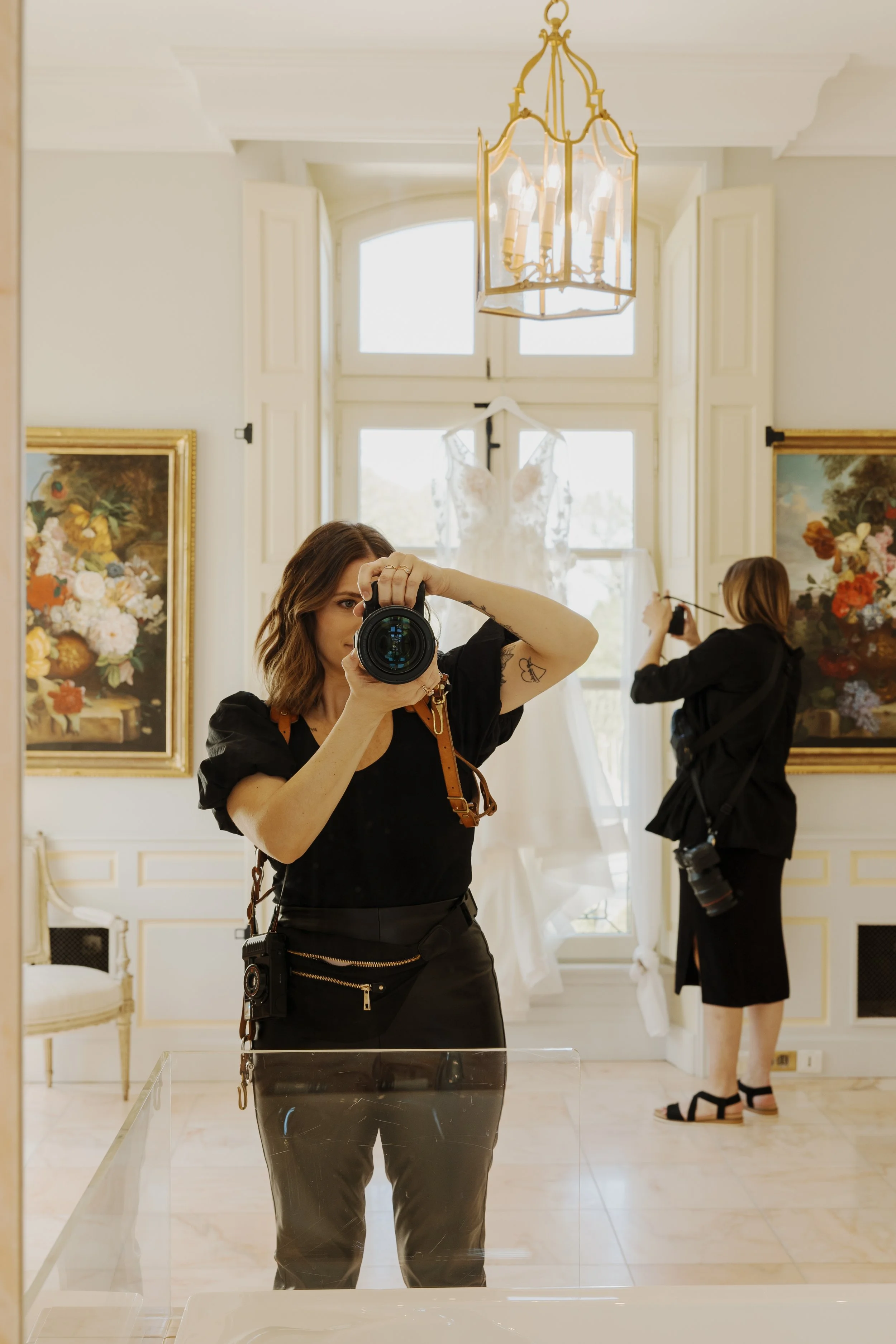 Grace Troutman taking photos of a white dress hanging by the window in an elegant room with framed floral paintings and chandelier.