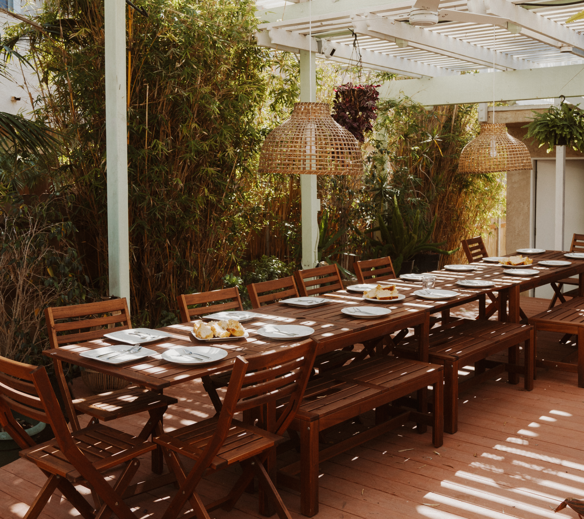 Outdoor dining area with a long wooden table set for dinner, surrounded by wooden chairs, with hanging wicker lamps and lush green plants on the side at Grace Troutman's educational photography retreat in Southern California.