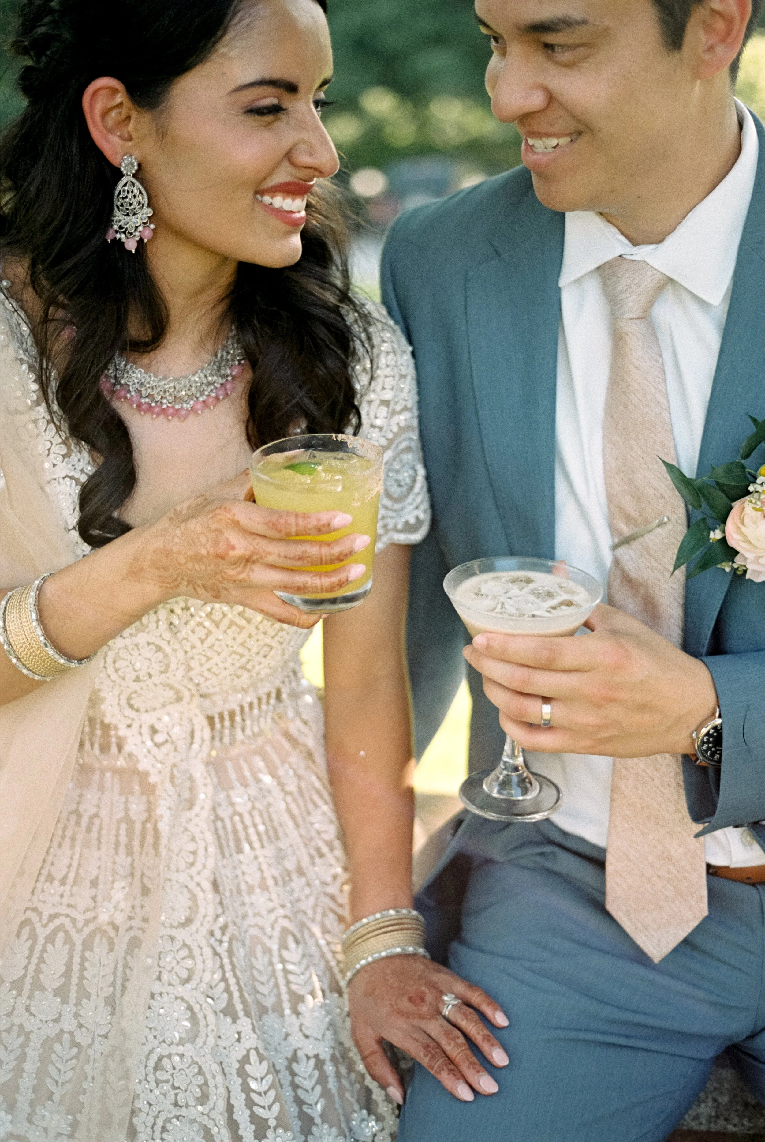 A woman and a man dressed in formal wedding attire are standing closely together, smiling and holding cocktails during a celebration.