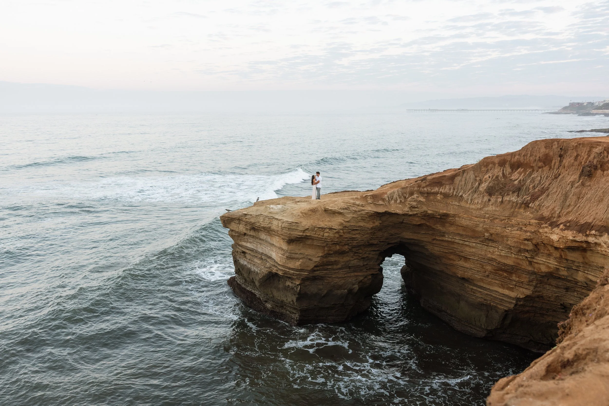 A couple embraces on a rocky cliff overlooking the ocean at Sunset Cliffs in San Diego California, with a natural arch formation in the foreground and a cloudy sky above.
