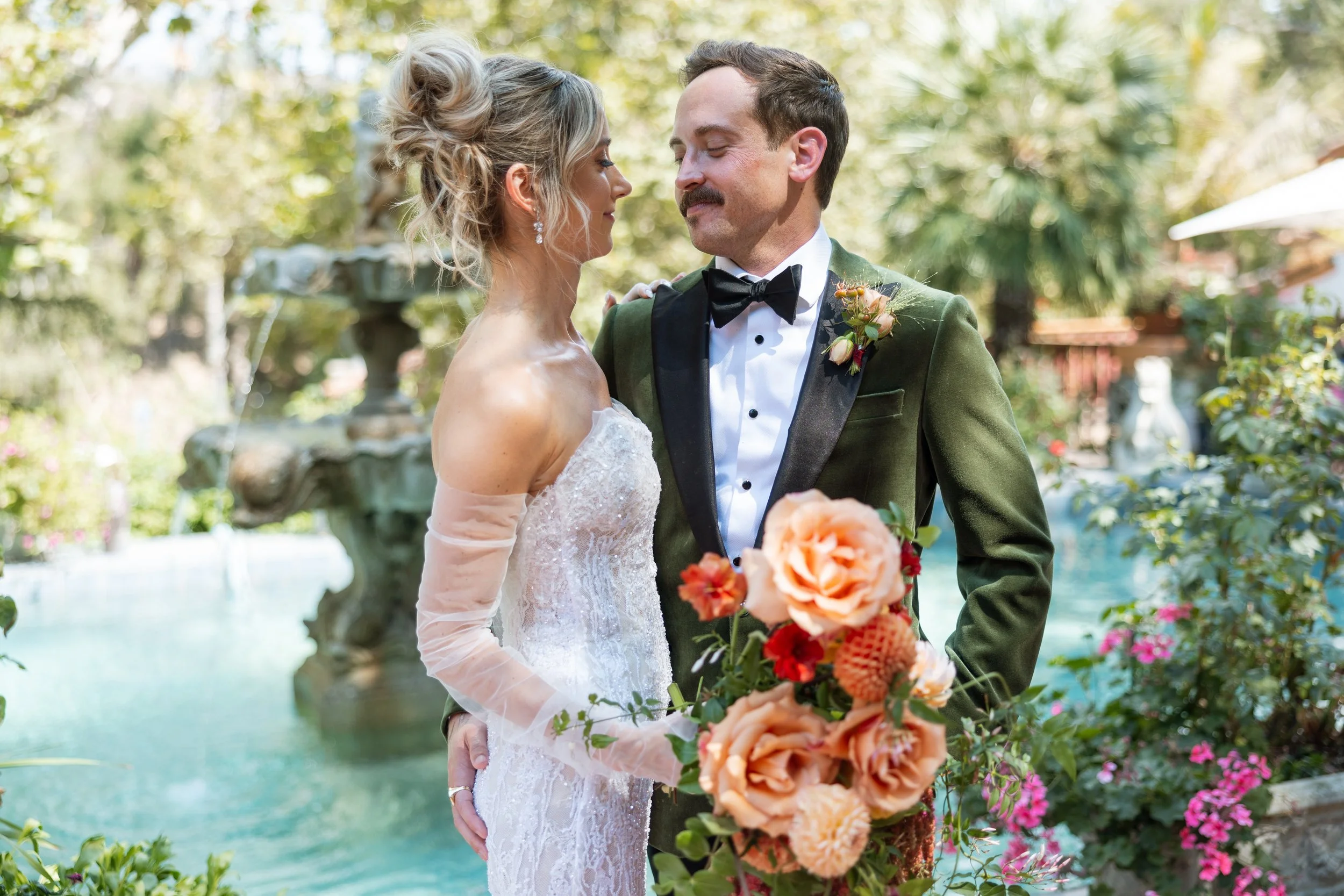 A bride and groom standing close together outdoors during a wedding, with a fountain and greenery in the background. The bride is wearing a white lace wedding dress and the groom is in a dark green tuxedo with a black bow tie, holding a bouquet of large peach and orange flowers.
