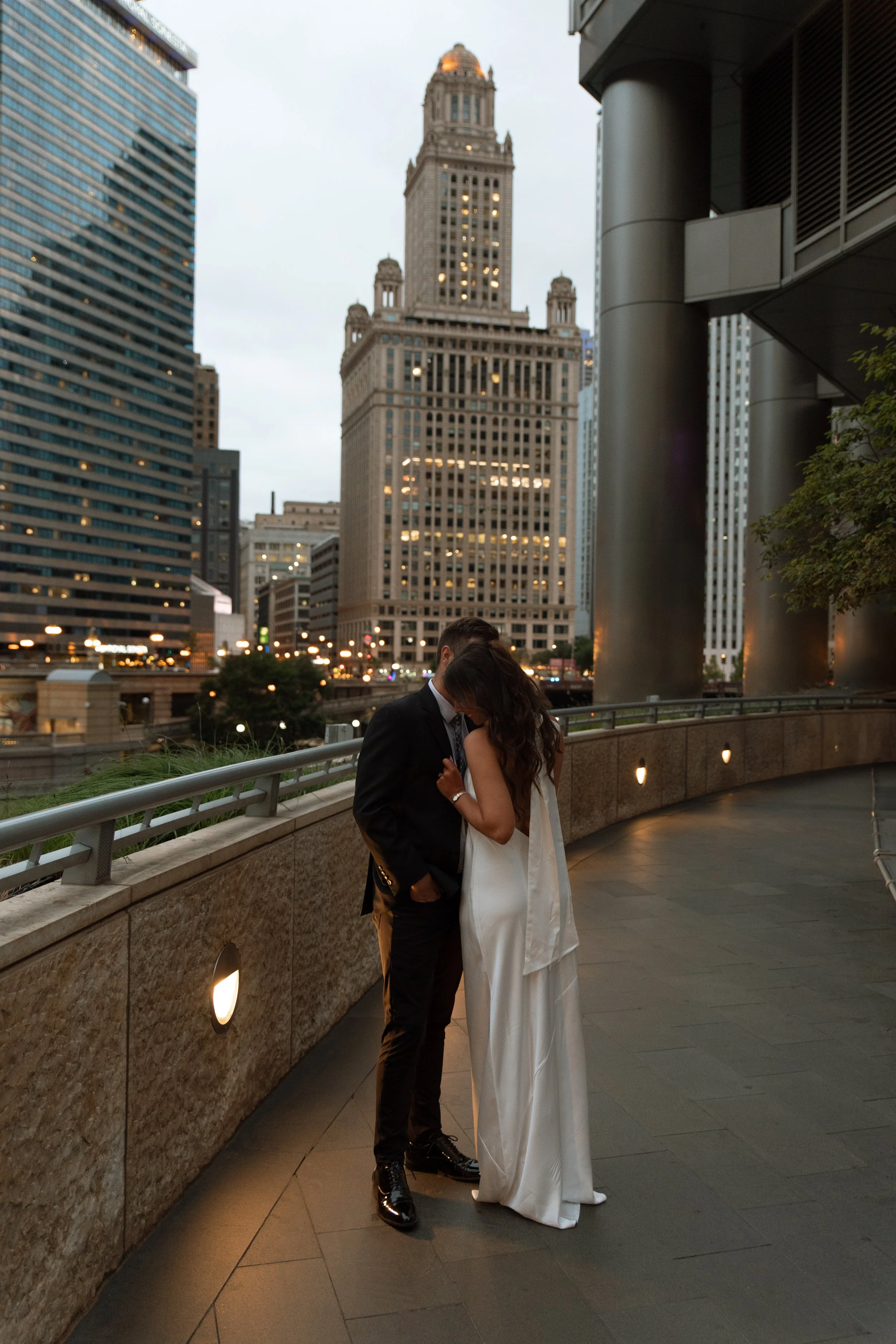 A couple dressed in formal wedding attire, sharing a romantic moment on an urban rooftop at dusk with city skyscrapers in the background.
