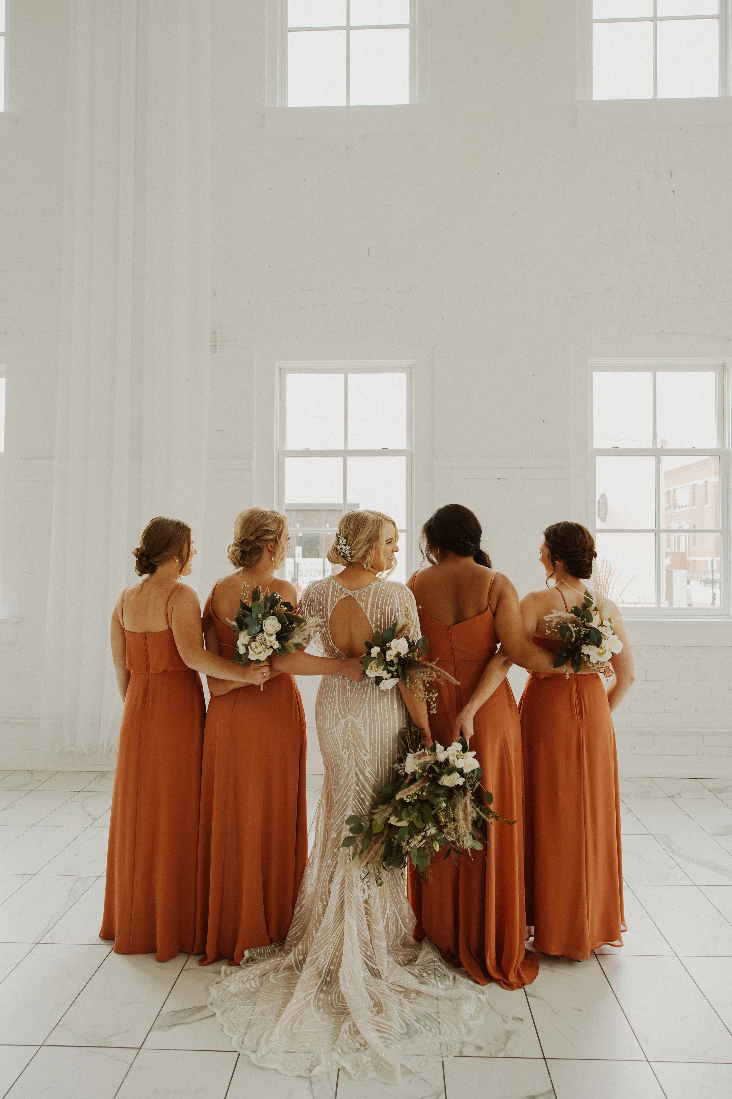 A bride and her four bridesmaids posing in a bright, white-walled room with large windows. The bride is wearing a white, beaded wedding gown, and the bridesmaids are dressed in matching rust-colored dresses, each holding a bouquet of white and green flowers.