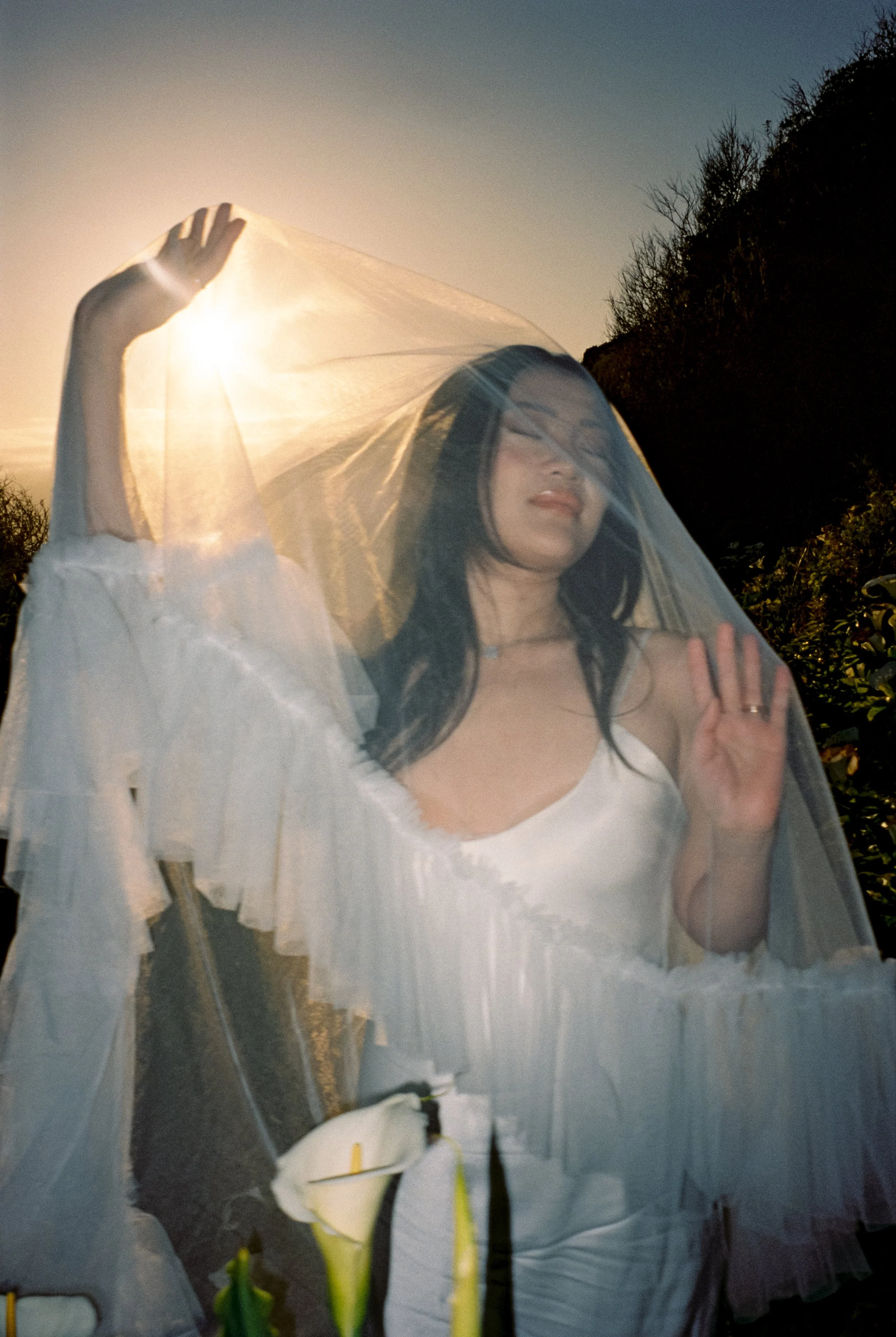 A woman in a white dress behind a sheer veil in an outdoor wedding during sunset.