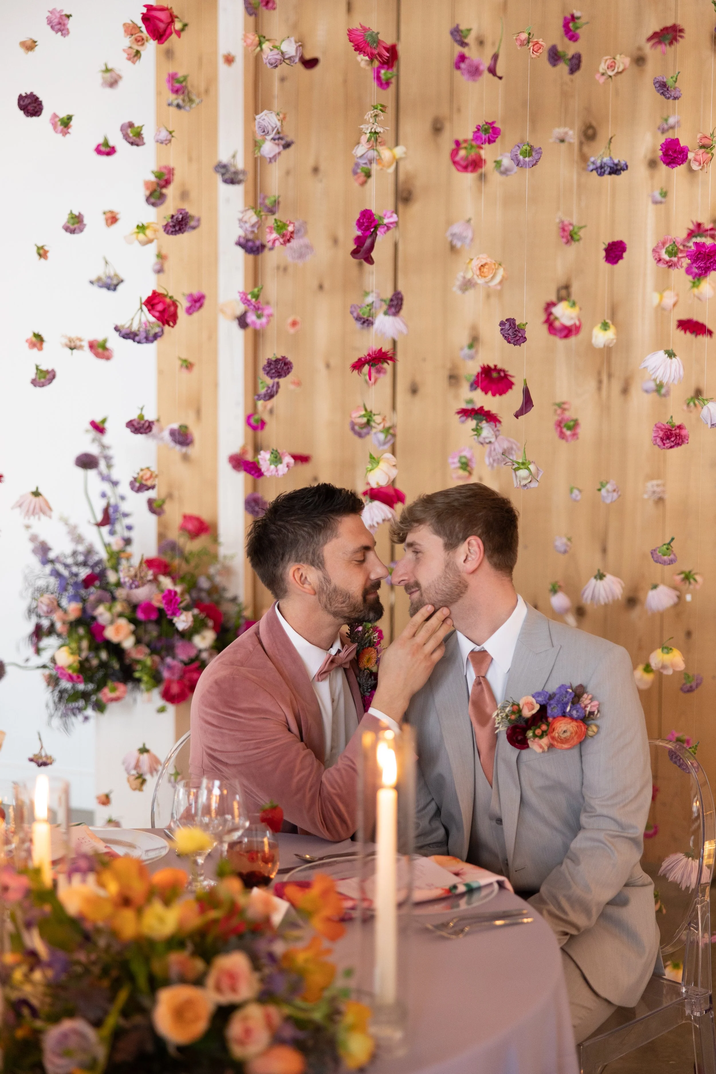 Two men, dressed in suits, are sitting close to each other at a wedding reception. One man is gently touching the other's face, and they are about to kiss. The background is decorated with suspended flowers and a floral arrangement on the table, creating a romantic atmosphere.