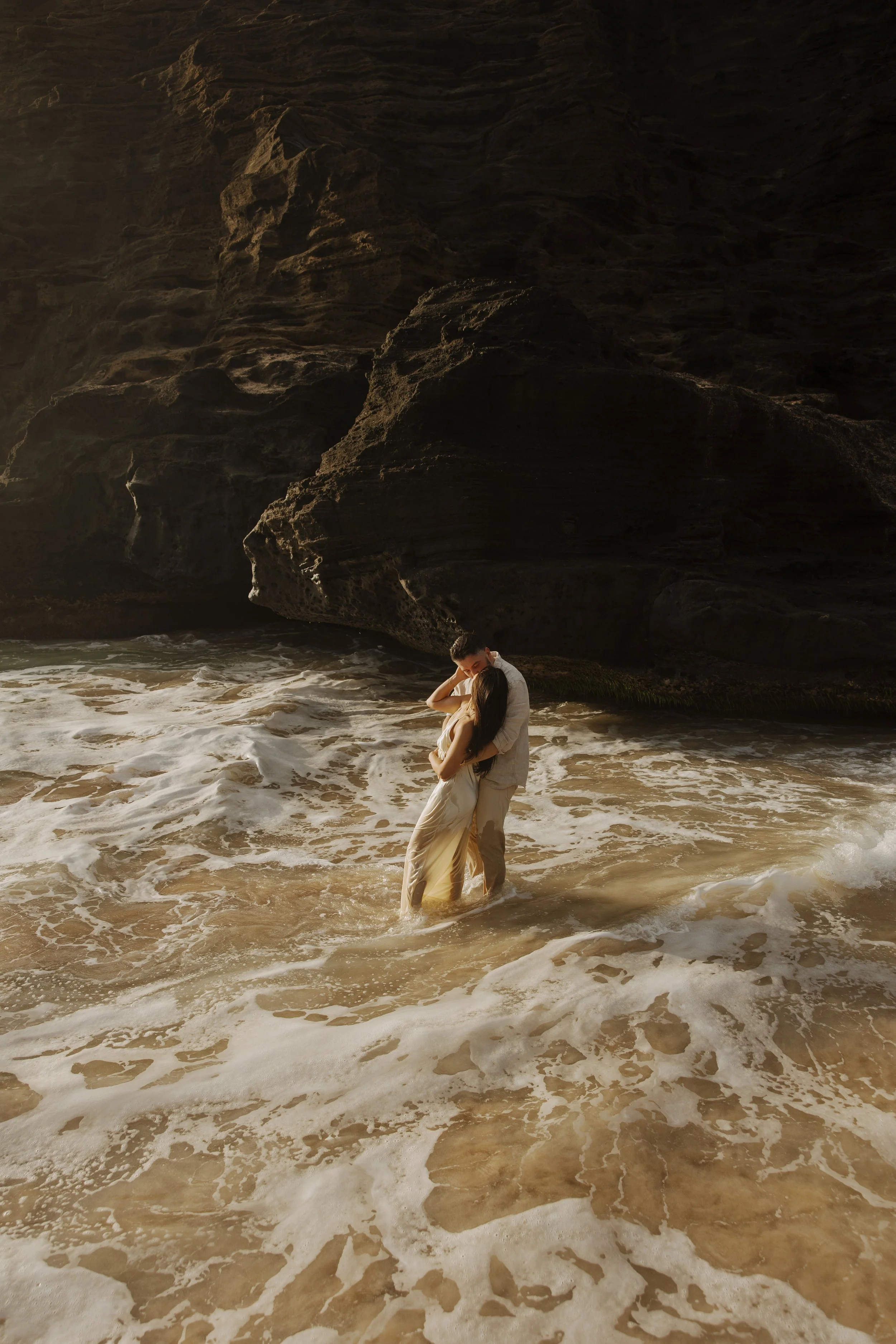 A couple dancing in the ocean waves at a Hawaiian beach with rocky cliffs in the background.