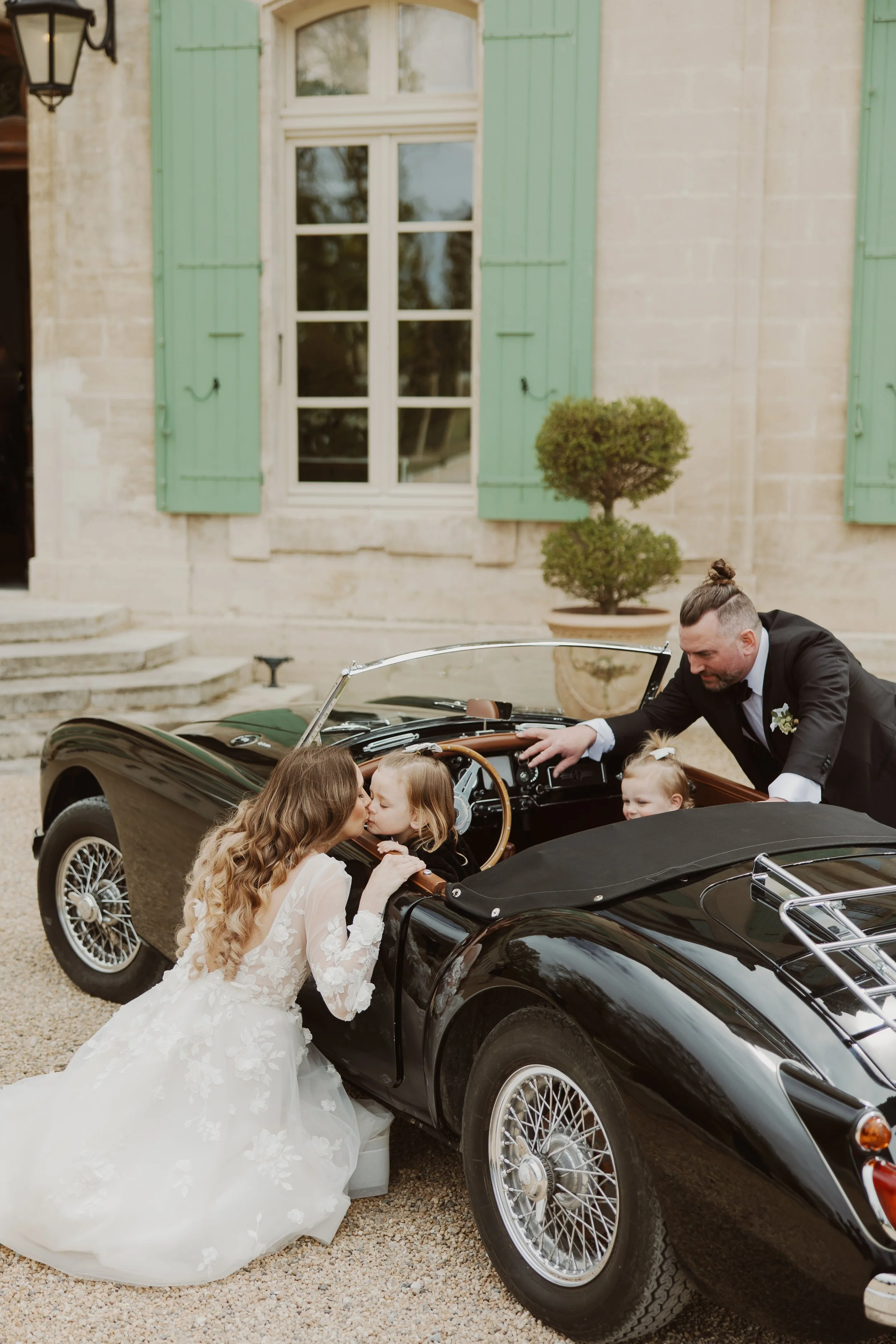 A bride and a groom with two young girls near a black vintage convertible car in front of a stone building with green shutters during a wedding celebration.