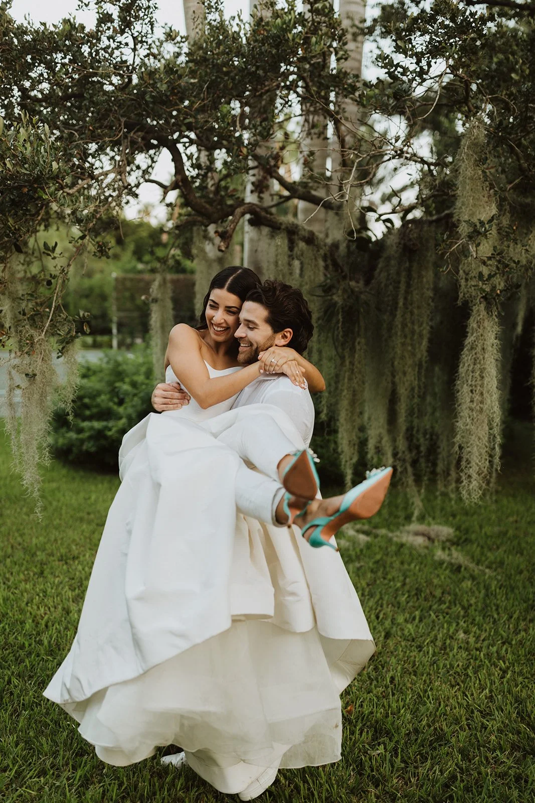 A couple is sitting on the grass in a garden, with the woman sitting on the man's lap. They are both smiling and embracing, with the woman hugging the man's neck. The woman is wearing a white dress, and the man is wearing a light-colored shirt and pants. They are surrounded by greenery and hanging moss from a tree above them.