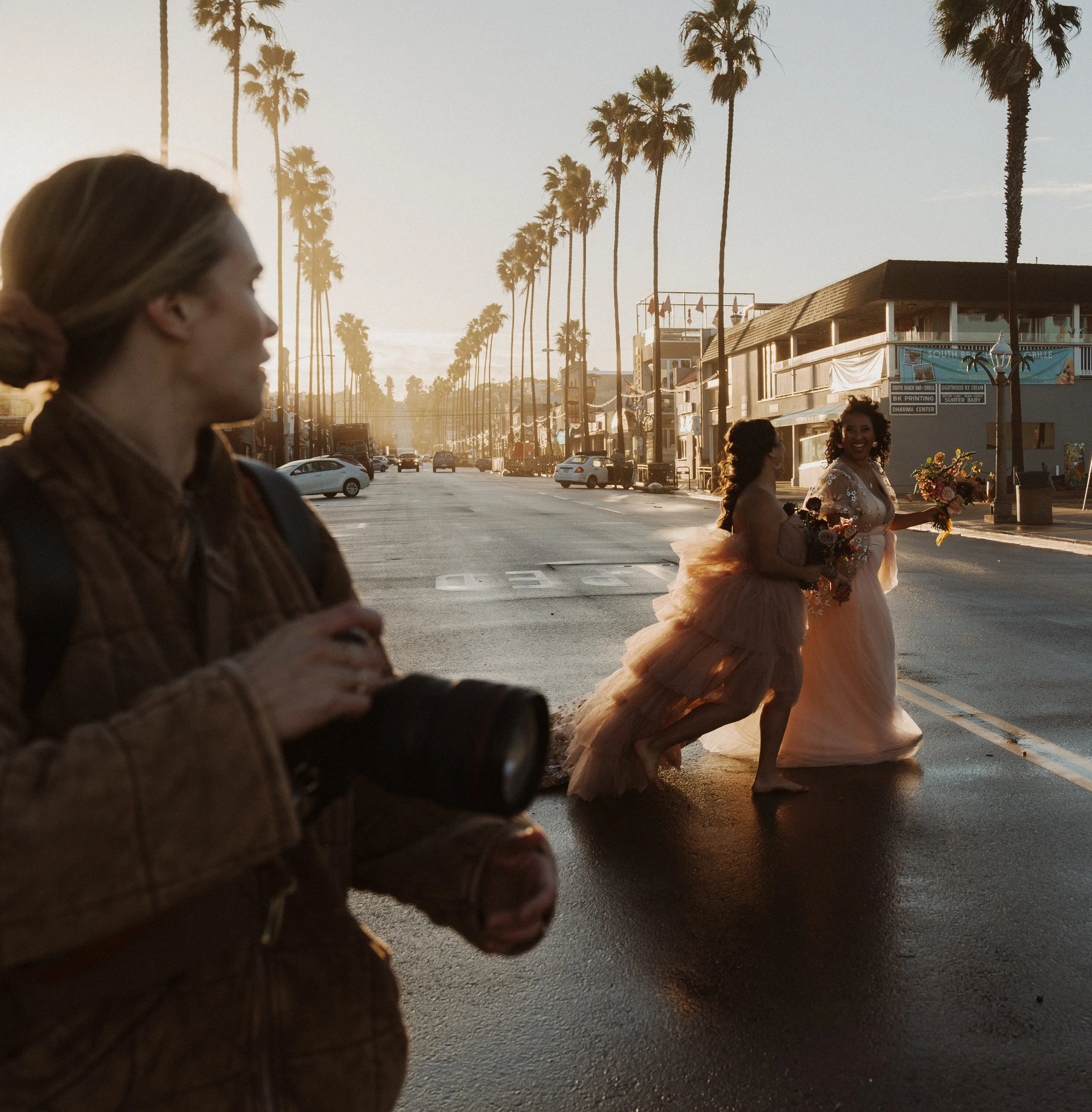 A lesbian couple in colorful textured wedding dresses walking on a street, one holding a bouquet, with palm trees and buildings in the background during sunset.