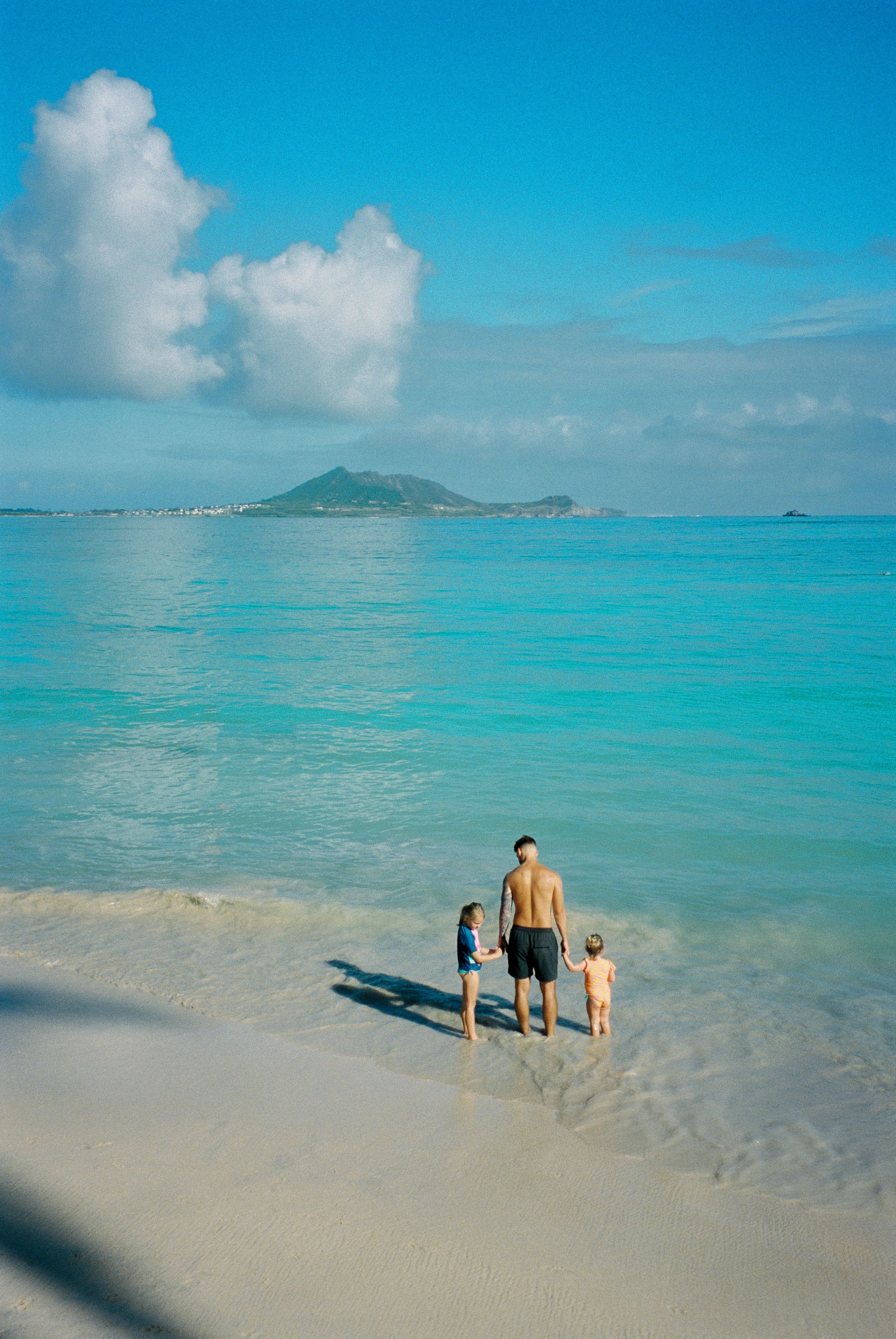 Grace Troutman's family at the beach in Hawaii, standing at the water's edge with ocean, clouds, and a distant mountain in the background.