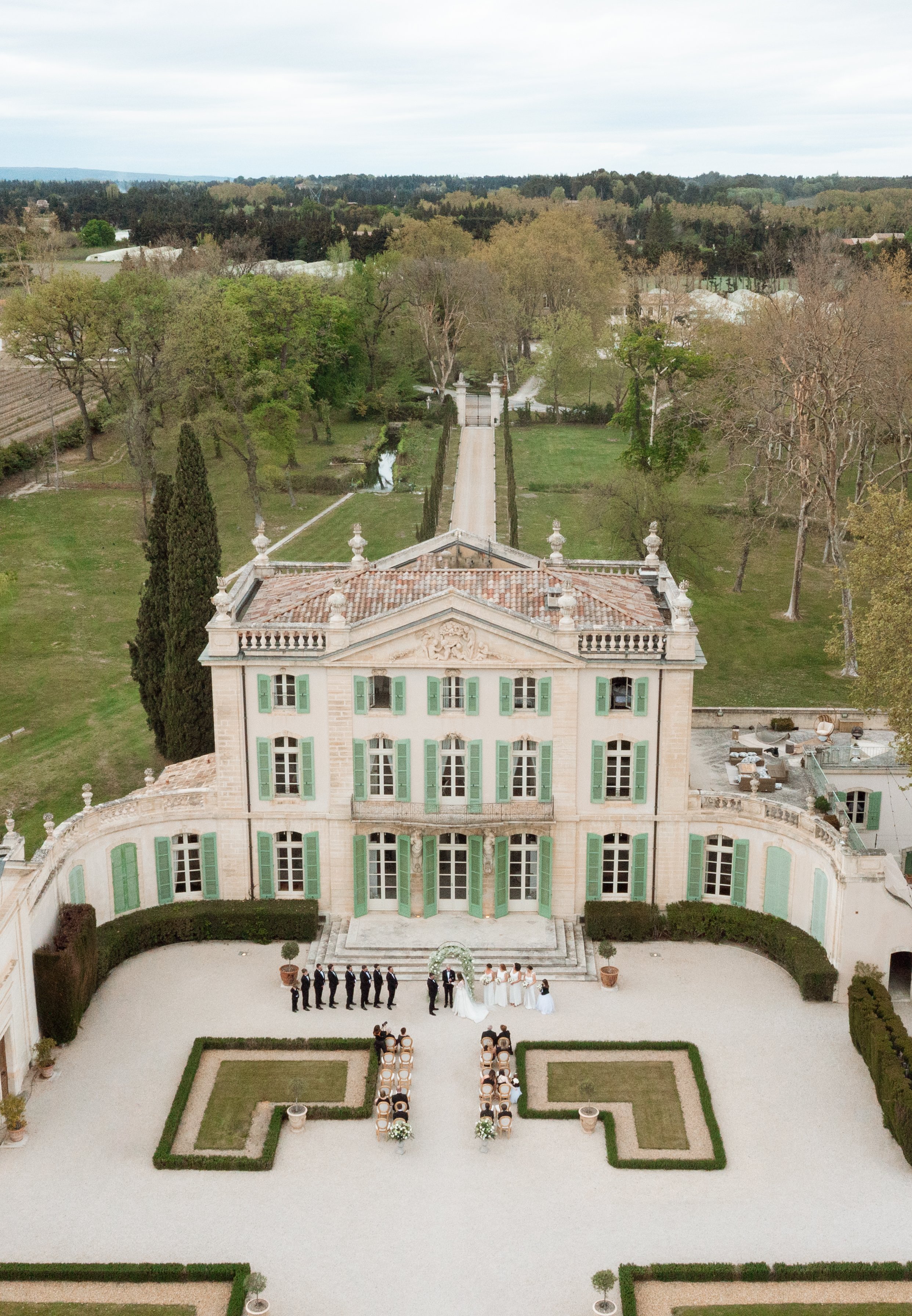 A wedding ceremony taking place outside a grand, historic mansion with green shutters, set in a large, landscaped garden with trees and a long driveway in the background.