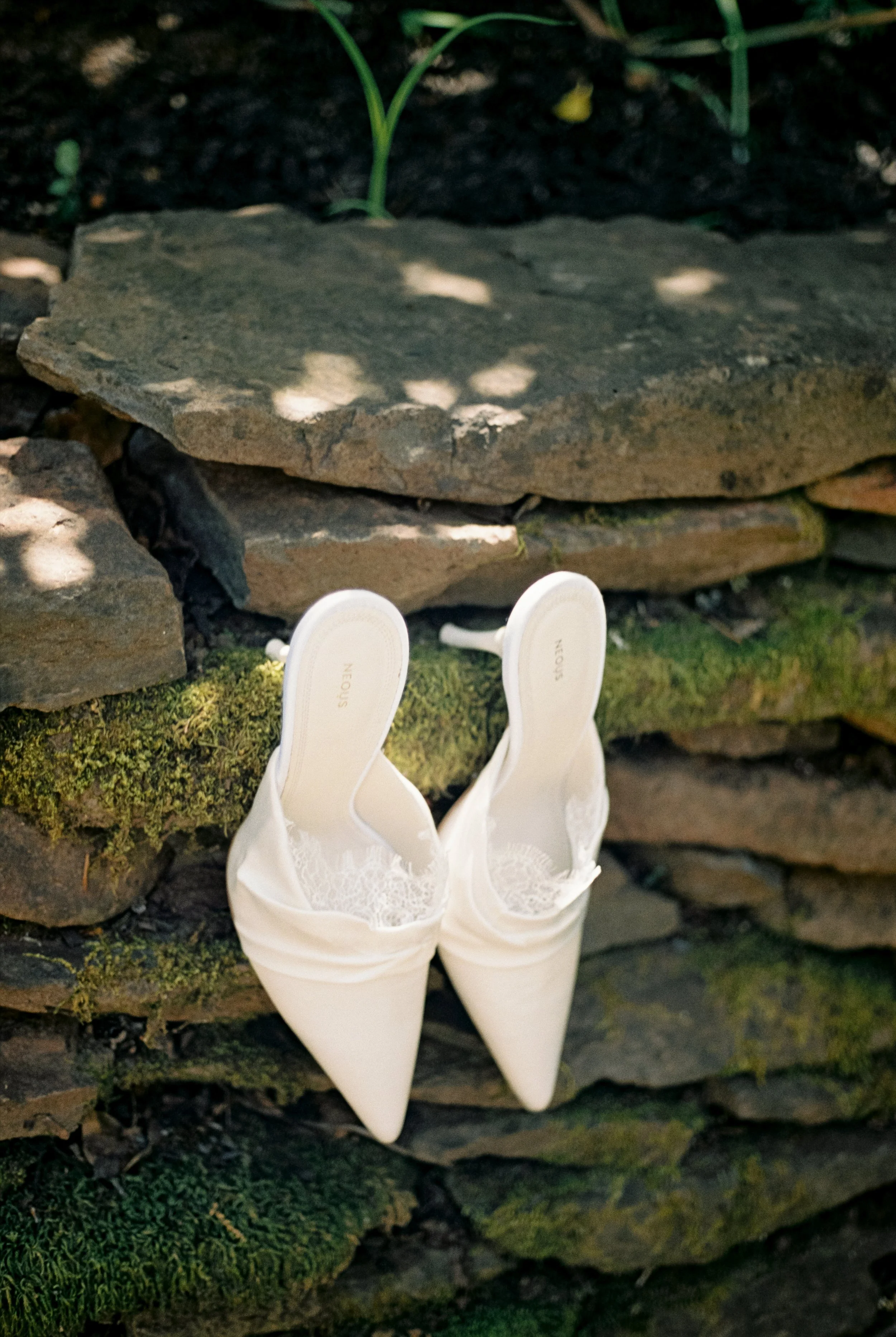 A bride's white high-heeled shoes placed on a moss-covered stone wall outdoors with sunlight filtering through trees.