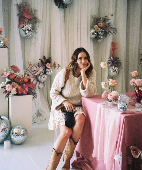 Grace Troutman sitting at a decorated table with pink and floral arrangements, smiling with her head resting on her hand, surrounded by pink and silver holiday-themed decor.