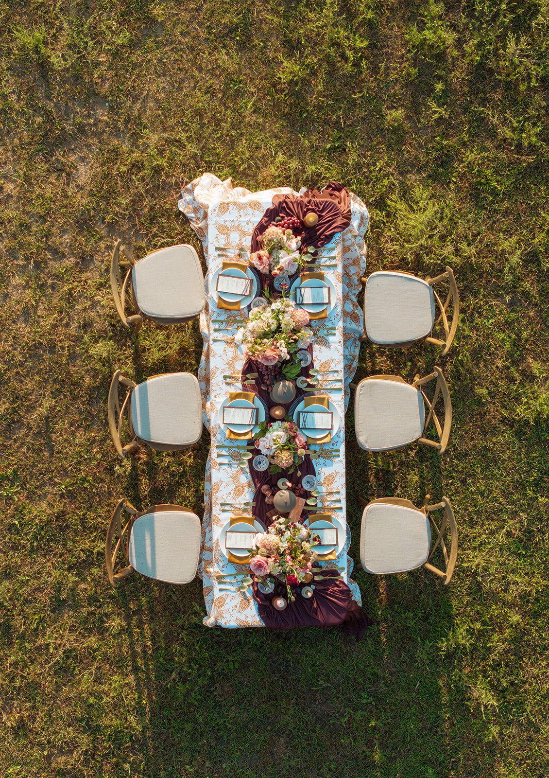 Overhead view of a decorated outdoor dining table with eight chairs on grass. The table features floral arrangements, candles, and elegant tableware, with a dark table runner.
