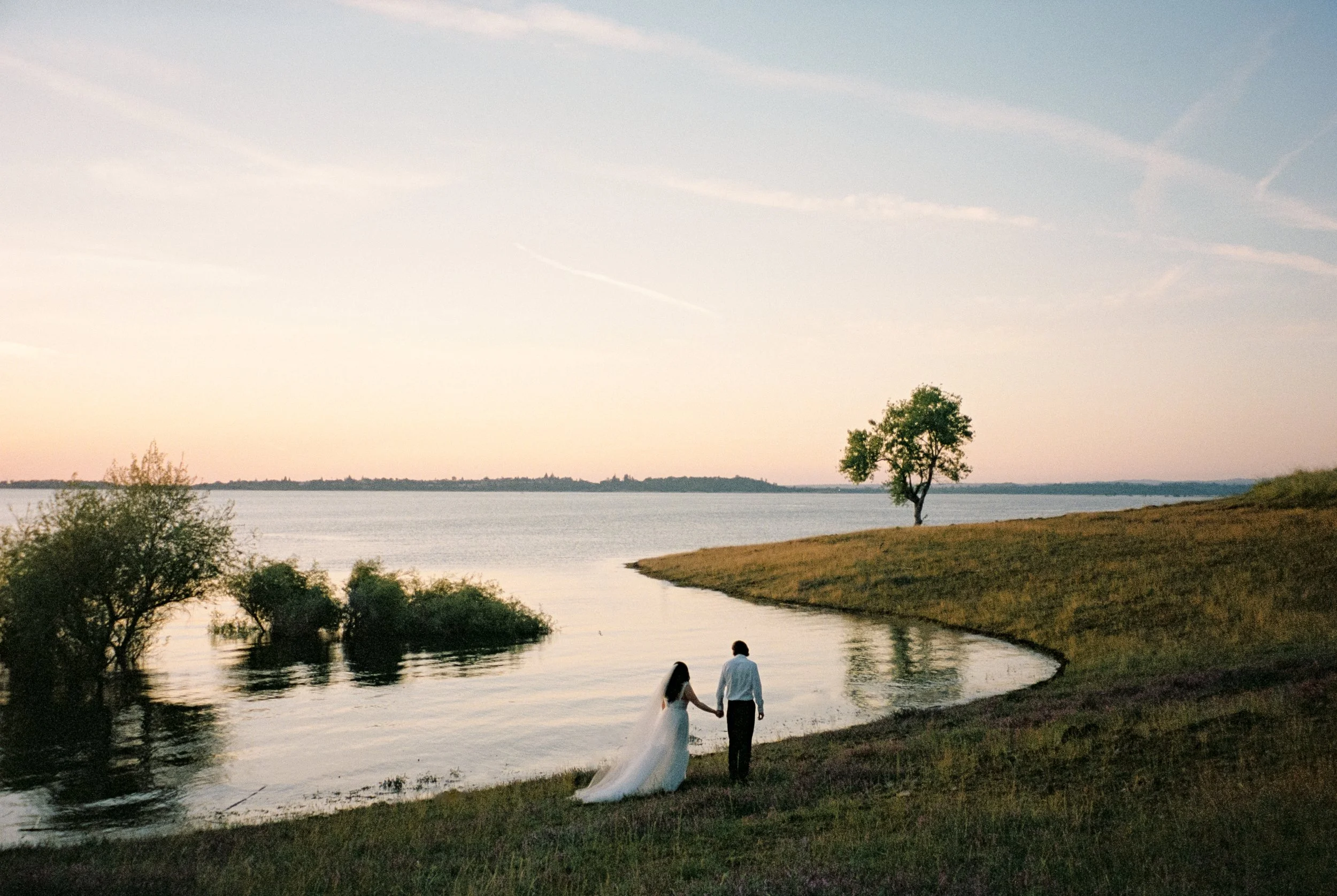 A couple walking hand-in-hand along a lakeshore at sunset, with a single tree on a grassy hill and water reflections.